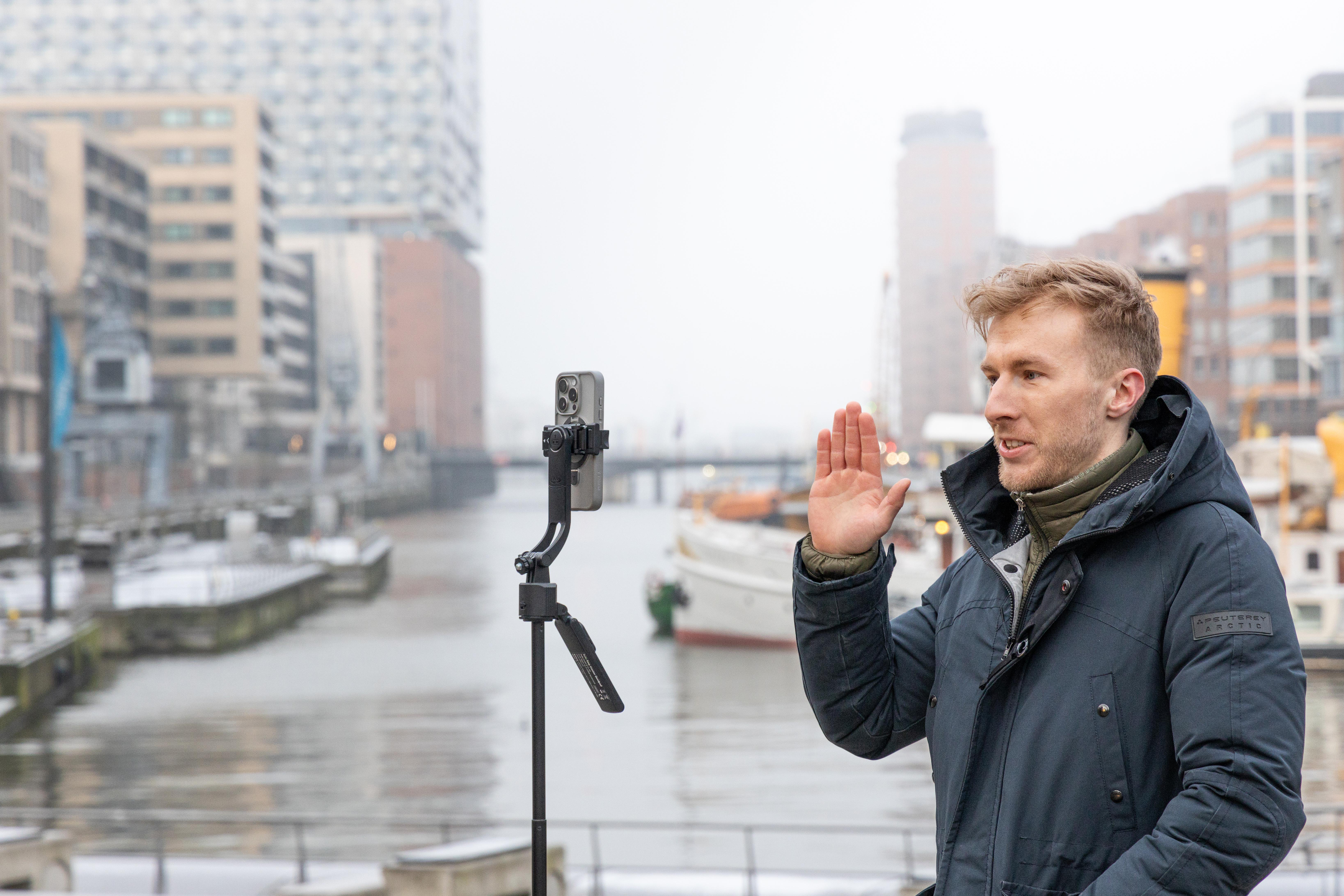 Mann mit erhobener Hand, der mit einem Telefon auf einem Stativ in der Nähe eines Gewässers filmt.