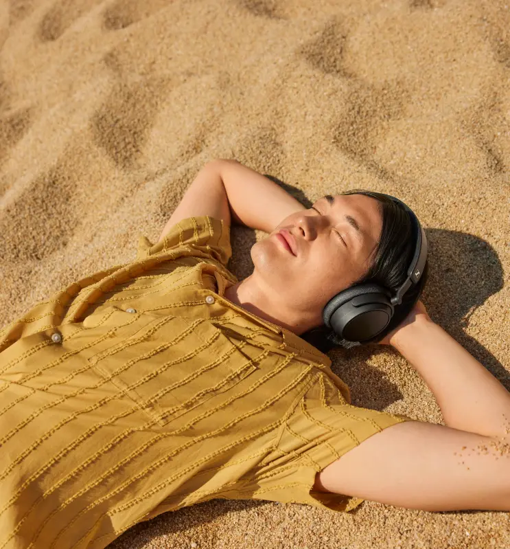 Eine Person liegt auf Sand, trägt Kopfhörer und ein gelbes Hemd, die Augen geschlossen. Sandiger Strandhintergrund.