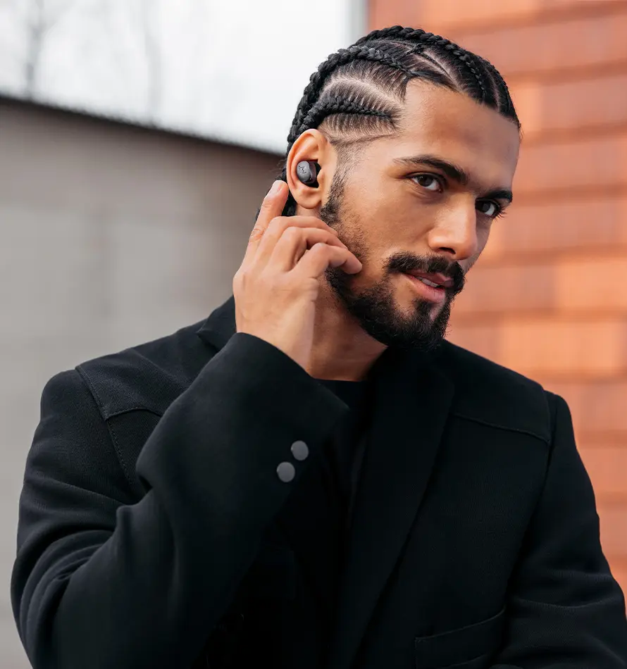 Un homme aux cheveux tressés et des écouteurs, portant une veste noire.