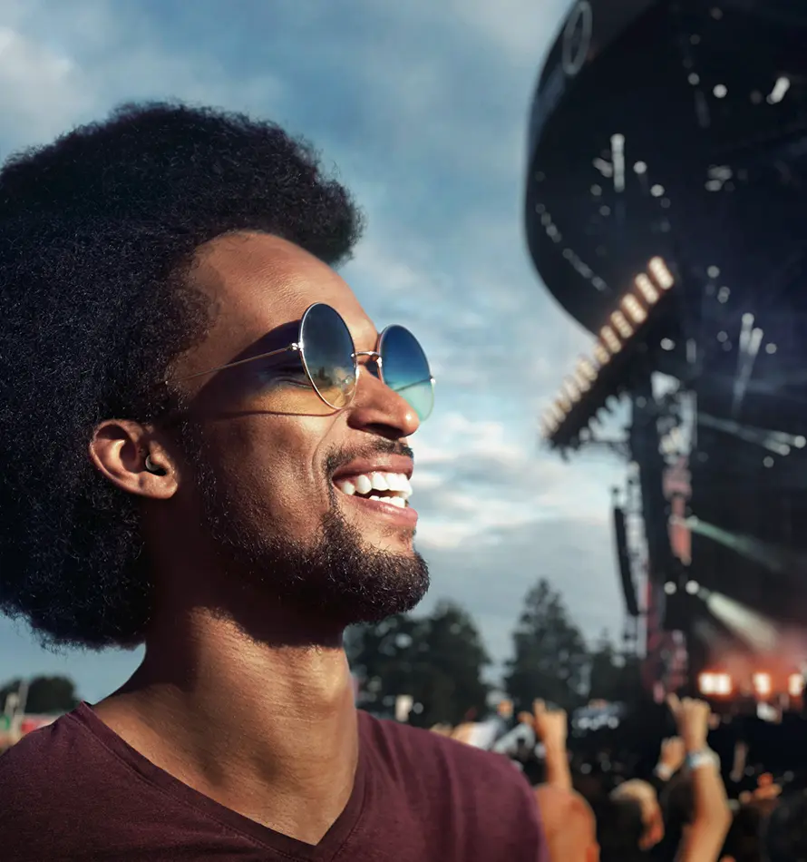 Homme avec des lunettes de soleil sourit à un concert, scène en arrière-plan, ciel ensoleillé et bleu.