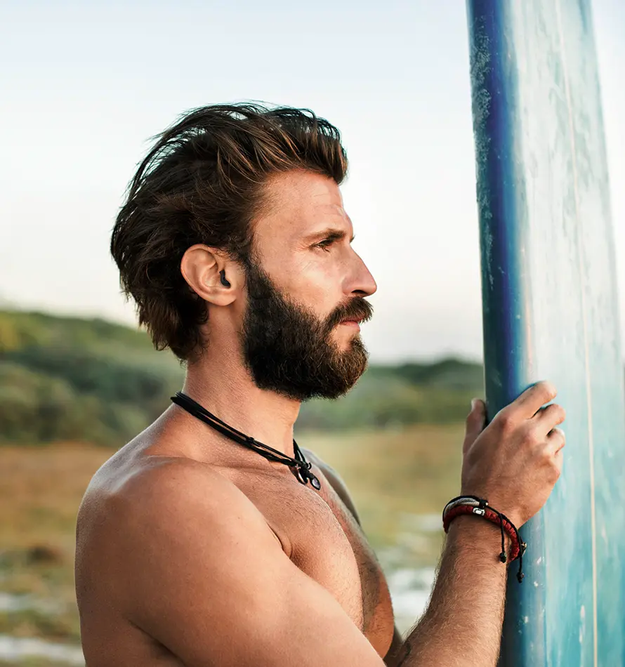 Un homme barbu tenant une planche de surf, regardant vers l'océan.