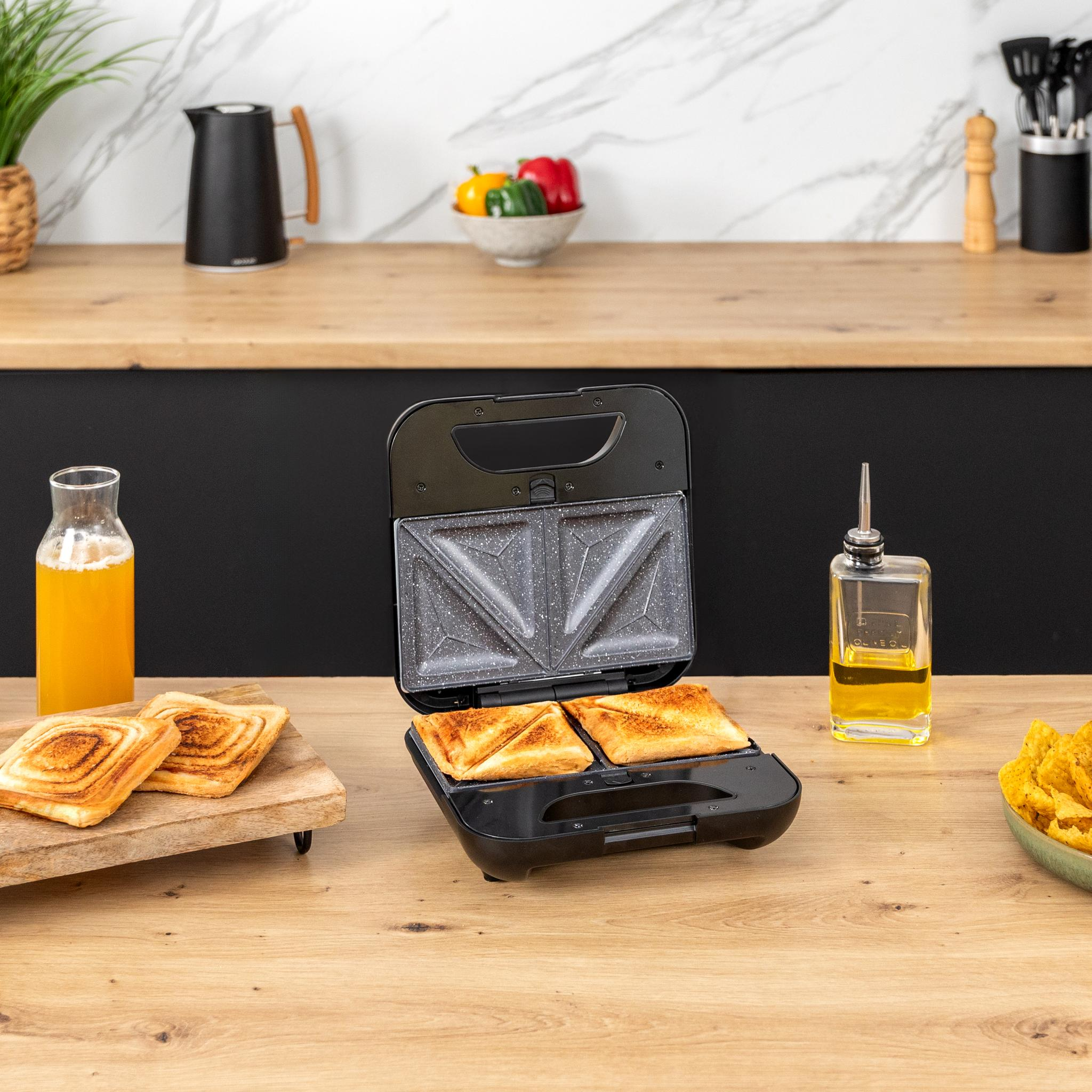 Una sandwichera negra con sándwiches tostados dentro. Una botella de zumo de naranja y patatas fritas están al lado.