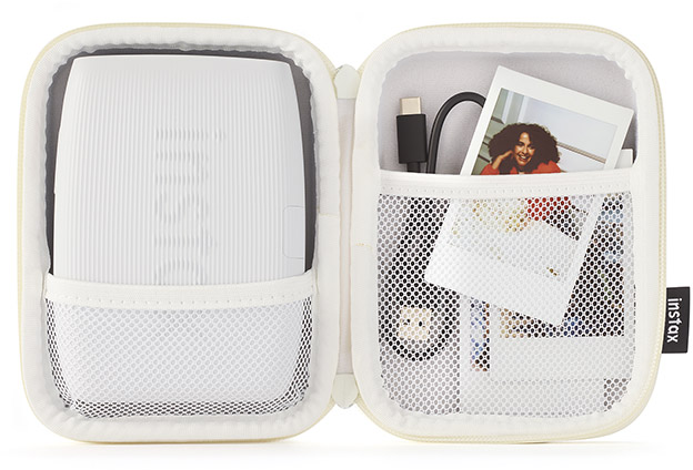 Fehér Instax nyomtató nyitott tokban, fotókkal és kábellel.