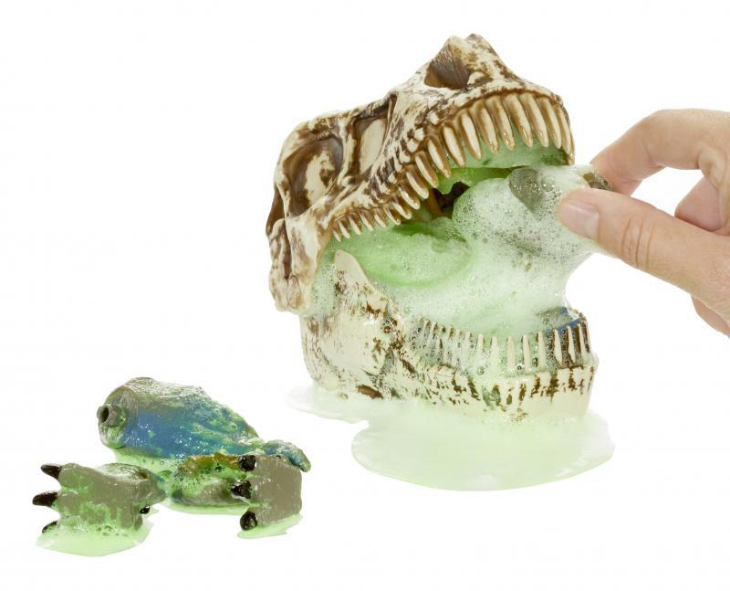 Dinosaurierschädel mit grünem Schaum, der herausspritzt. Eine Hand berührt ihn und ein kleiner Dino-Fuß ist in der Nähe.
