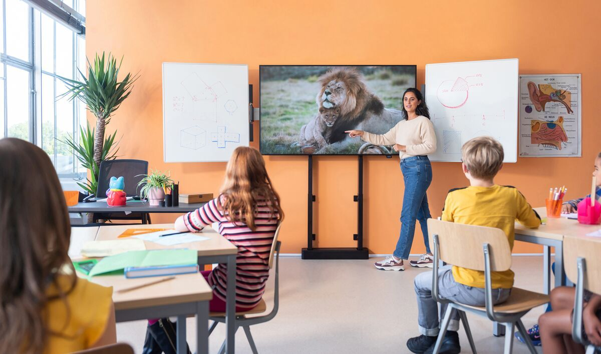 Klassenzimmer mit einer Lehrerin, die auf ein Bild eines Löwen zeigt, und Schülern an Schreibtischen.