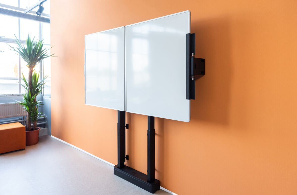 Zwei Whiteboards auf Ständern vor einer orangefarbenen Wand, mit einer Pflanze und einem orangefarbenen Sitz im Raum.