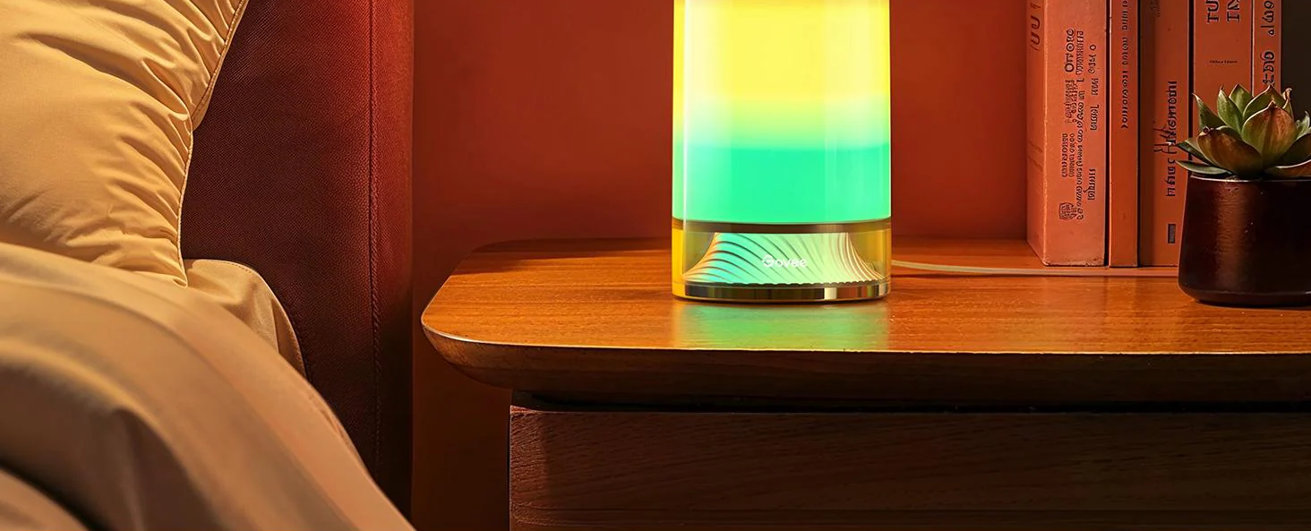 Comodino con una lampada, libri e una piccola pianta. La lampada ha sfumature di colore.