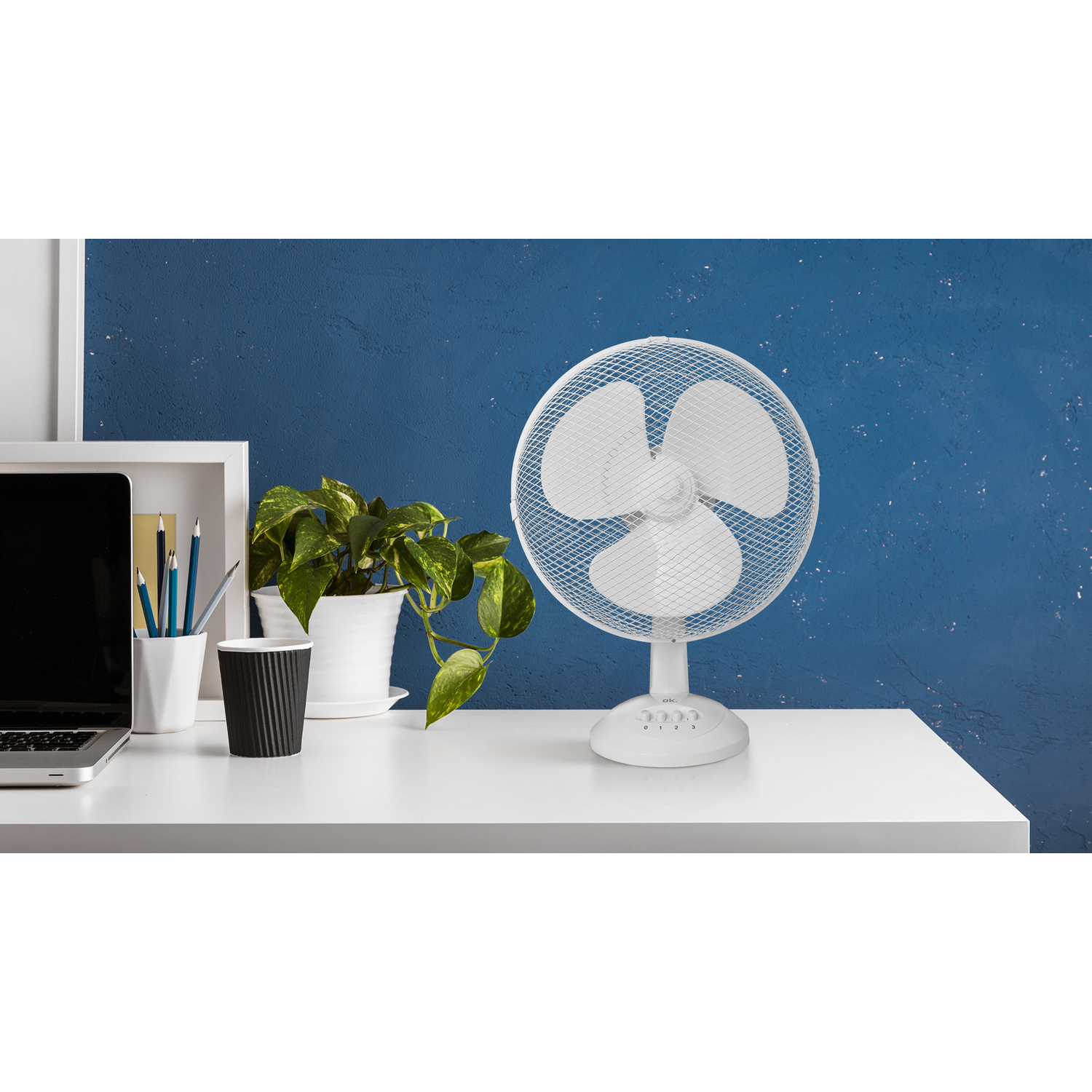 White desk fan with three blades. On a white desk with a laptop, pen holder, and plant.
