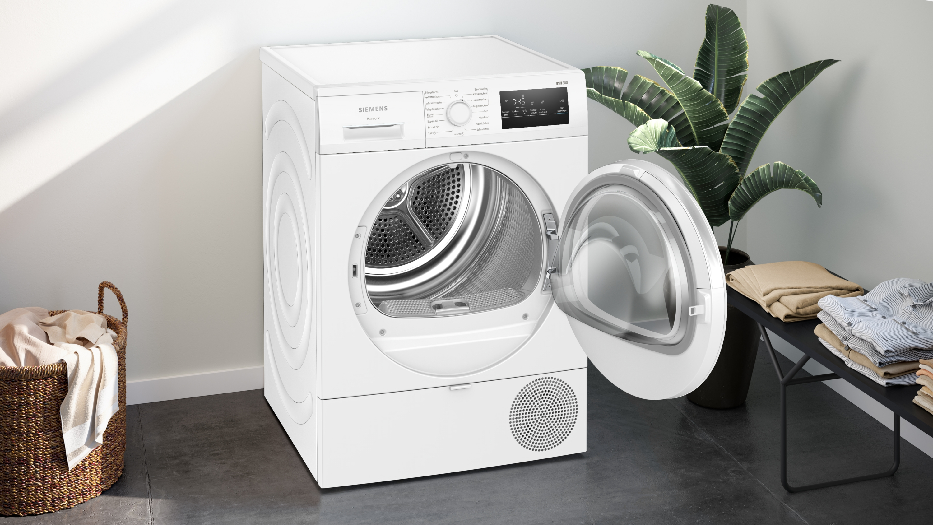 Ein weißer Siemens-Trockner mit geöffneter Tür, eine Pflanze, ein Wäschekorb und ein Tisch mit gefalteten Kleidern.