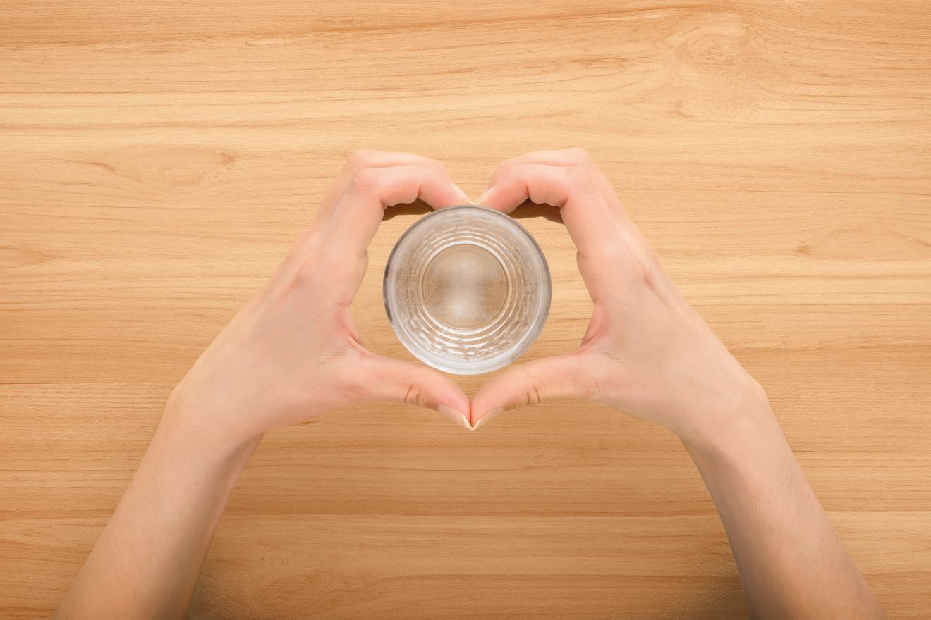 Hände bilden Herzform um ein Glas Wasser auf Holzoberfläche.