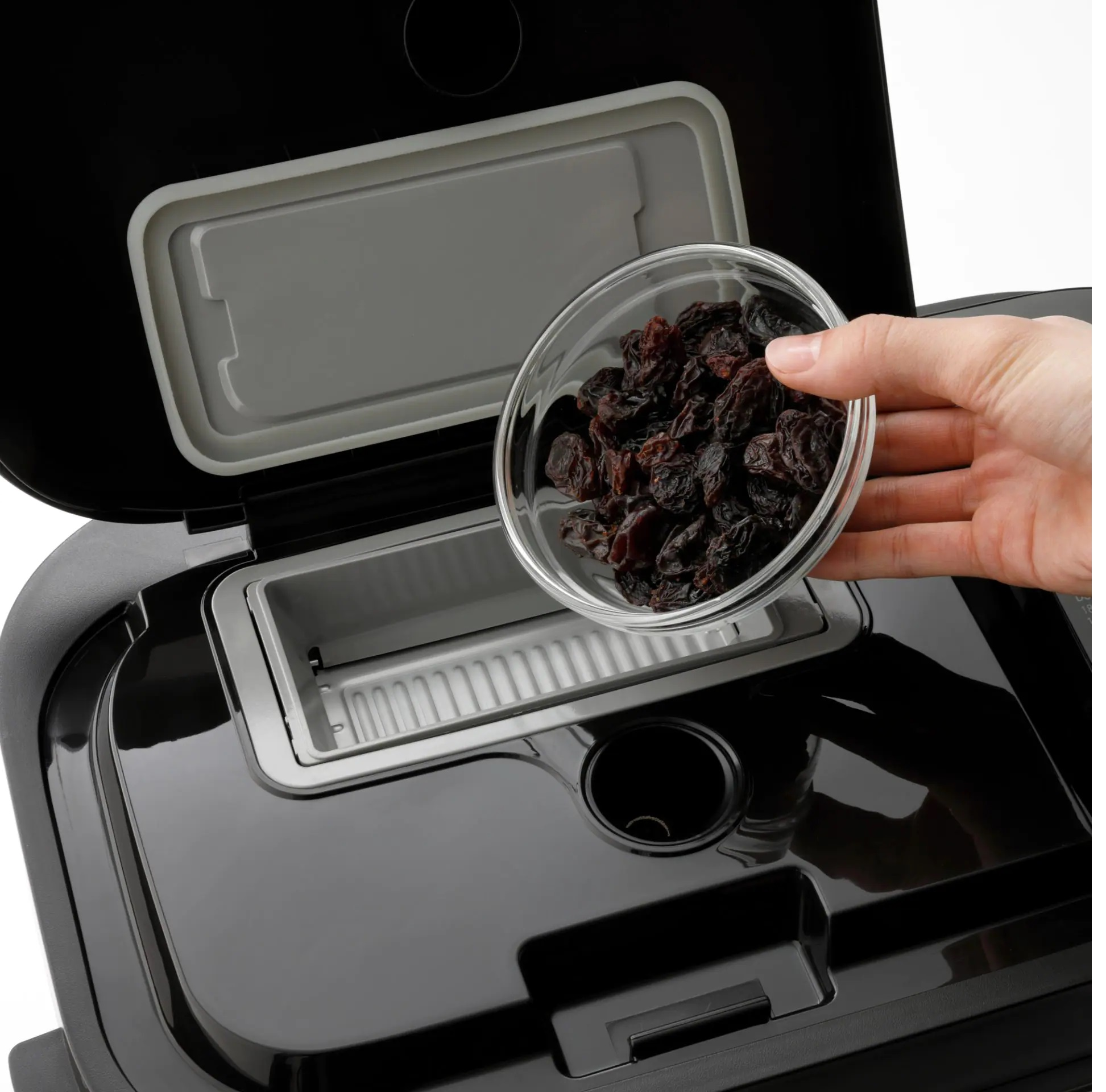 Czarna maszyna do chleba z otwartą pokrywą. Ręka wsypująca rodzynki ze szklanej miski.