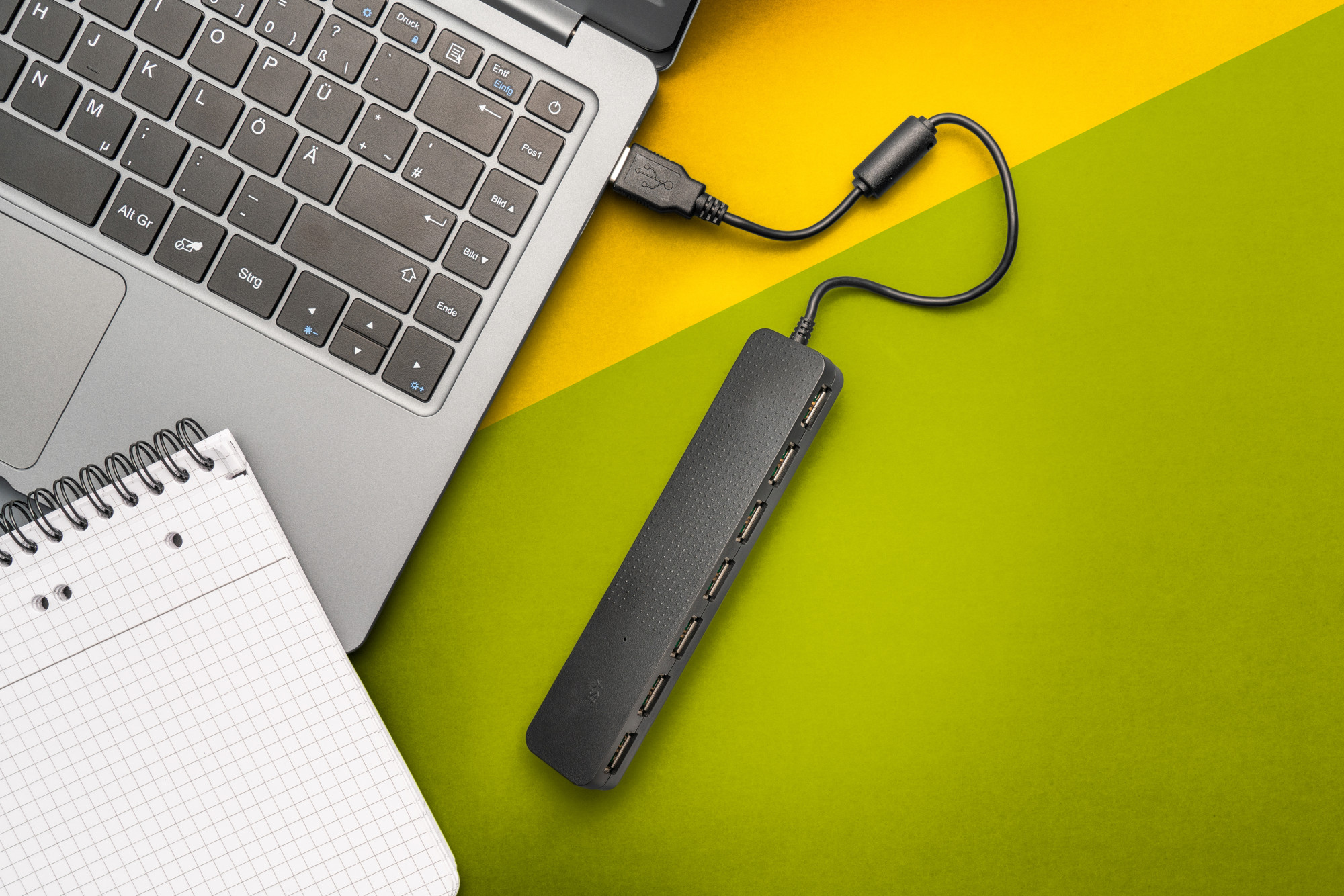 Laptop z hubem USB i notatnikiem na zielono-żółtej powierzchni. Widoczny podłączony kabel USB.