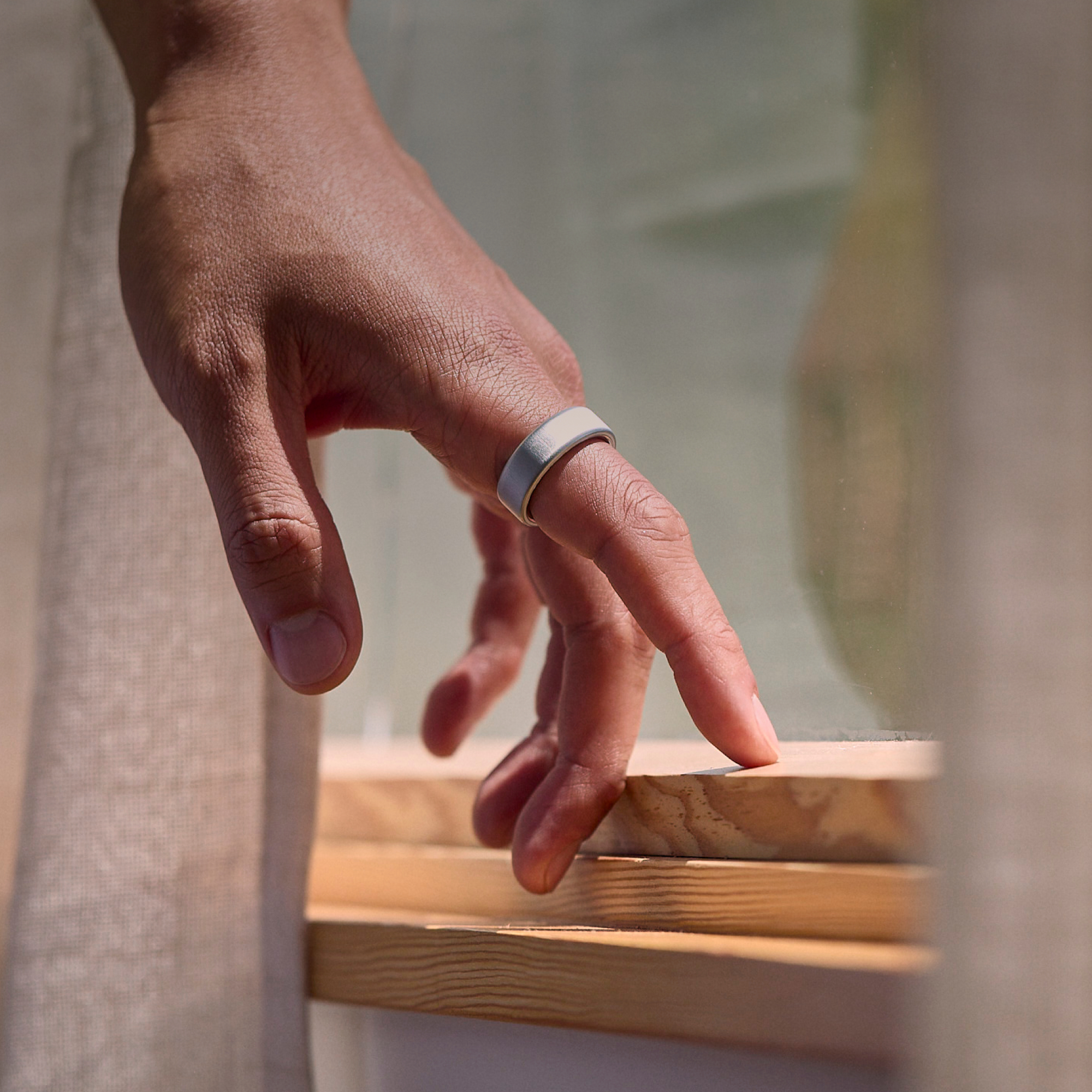 Eine Hand, die einen silbernen Oura-Ring an einem Finger trägt, ruht auf einer Holzoberfläche, in der Nähe eines Fensters.