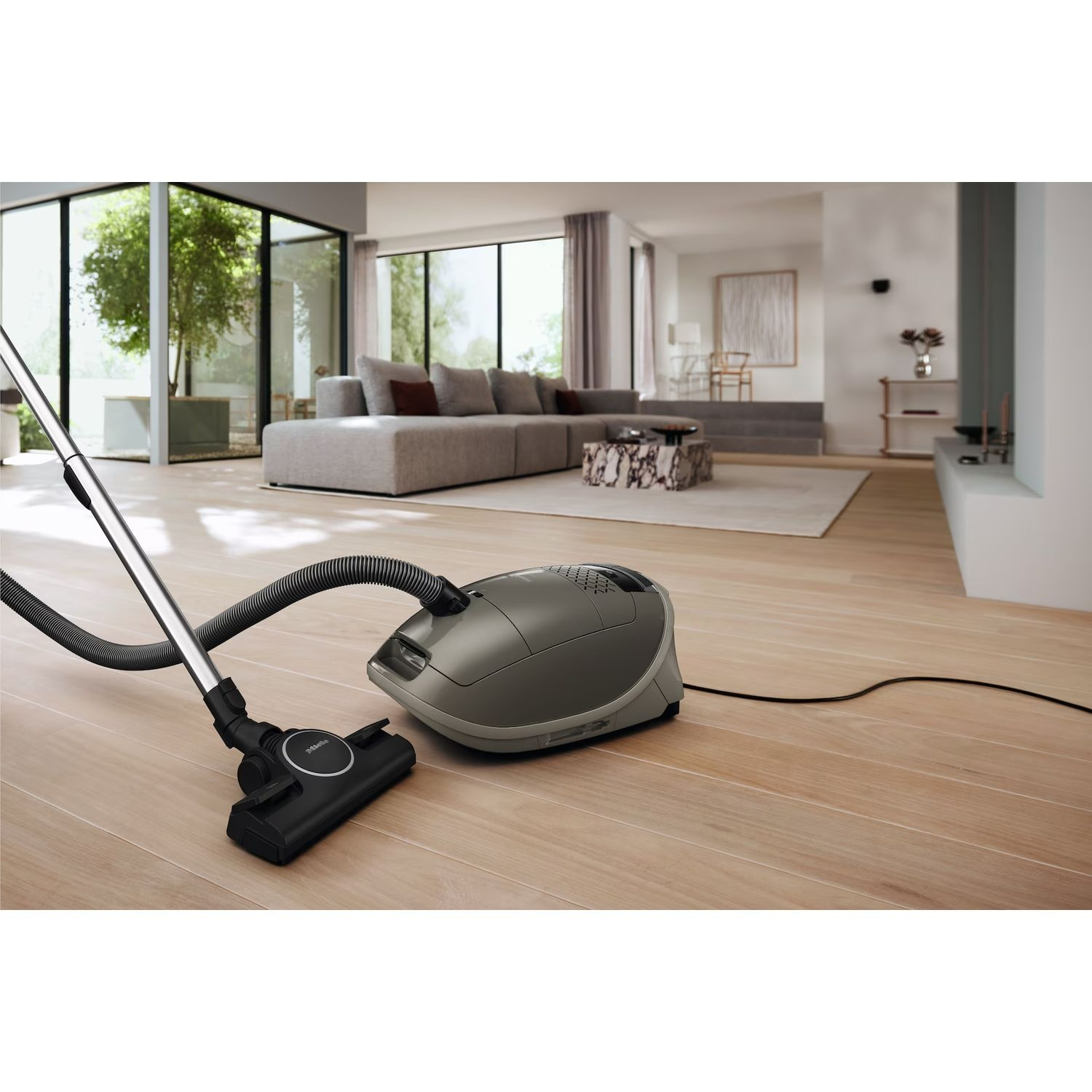 Aspirateur sur un plancher en bois, orienté vers la caméra, avec une fenêtre en arrière-plan.