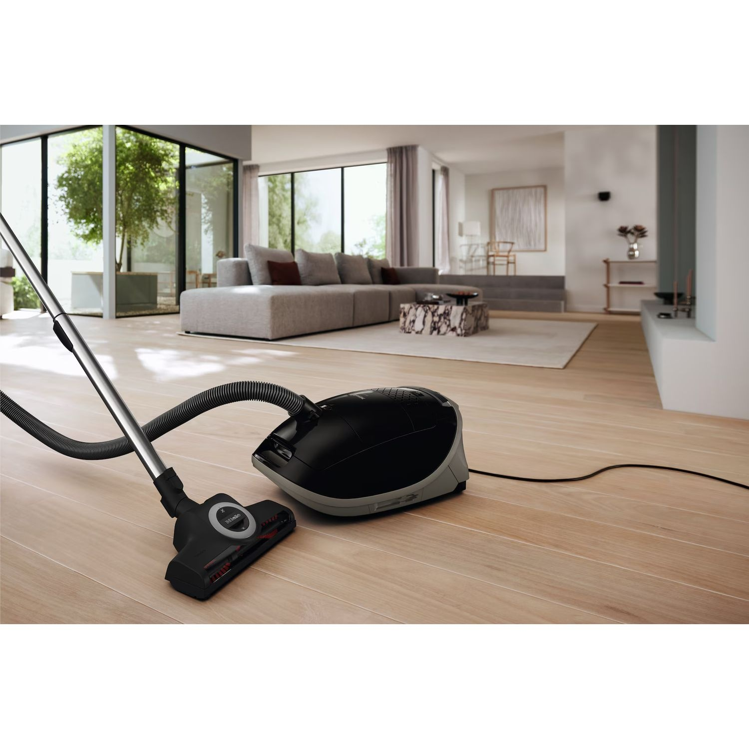 Un aspirateur noir et gris sur un plancher en bois, avec une fenêtre en arrière-plan.