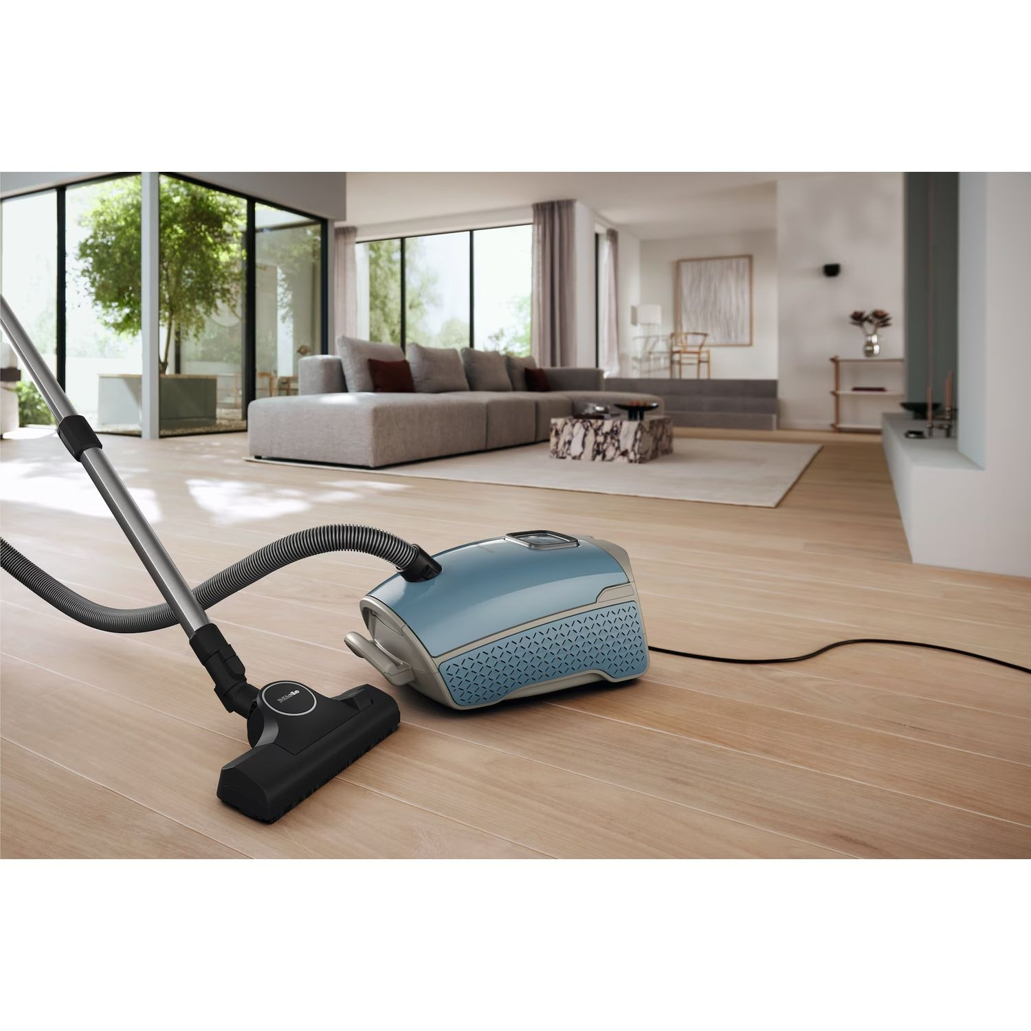 A blue vacuum cleaner on a wooden floor, hose and head visible. Brightly lit, clean interior.
