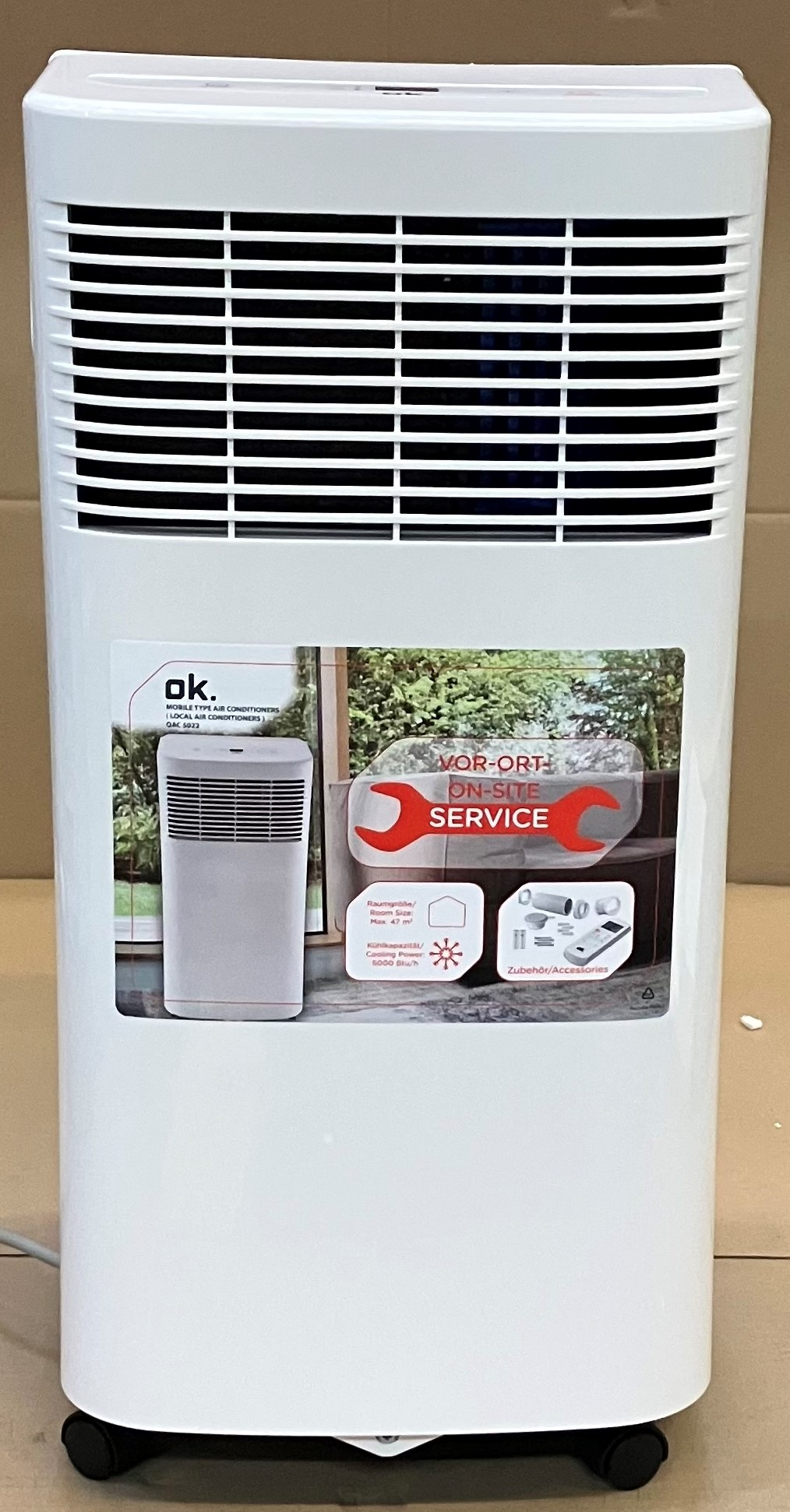 Biały klimatyzator z etykietą. Ma czarne koła. Tłem jest jasnobrązowa ściana.