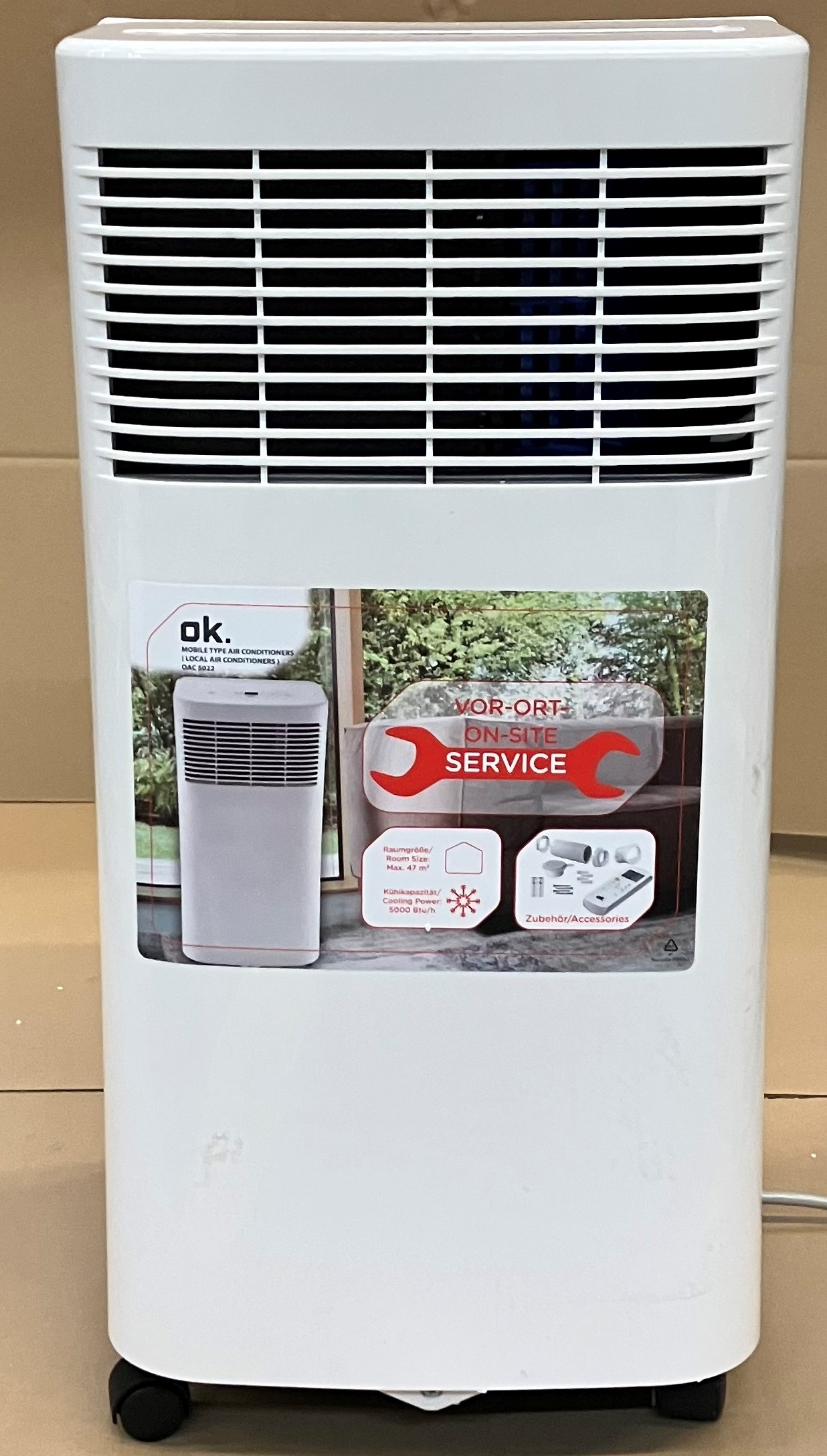 Biały klimatyzator z otworami wentylacyjnymi i naklejką. Urządzenie jest na kółkach. Białe tło.