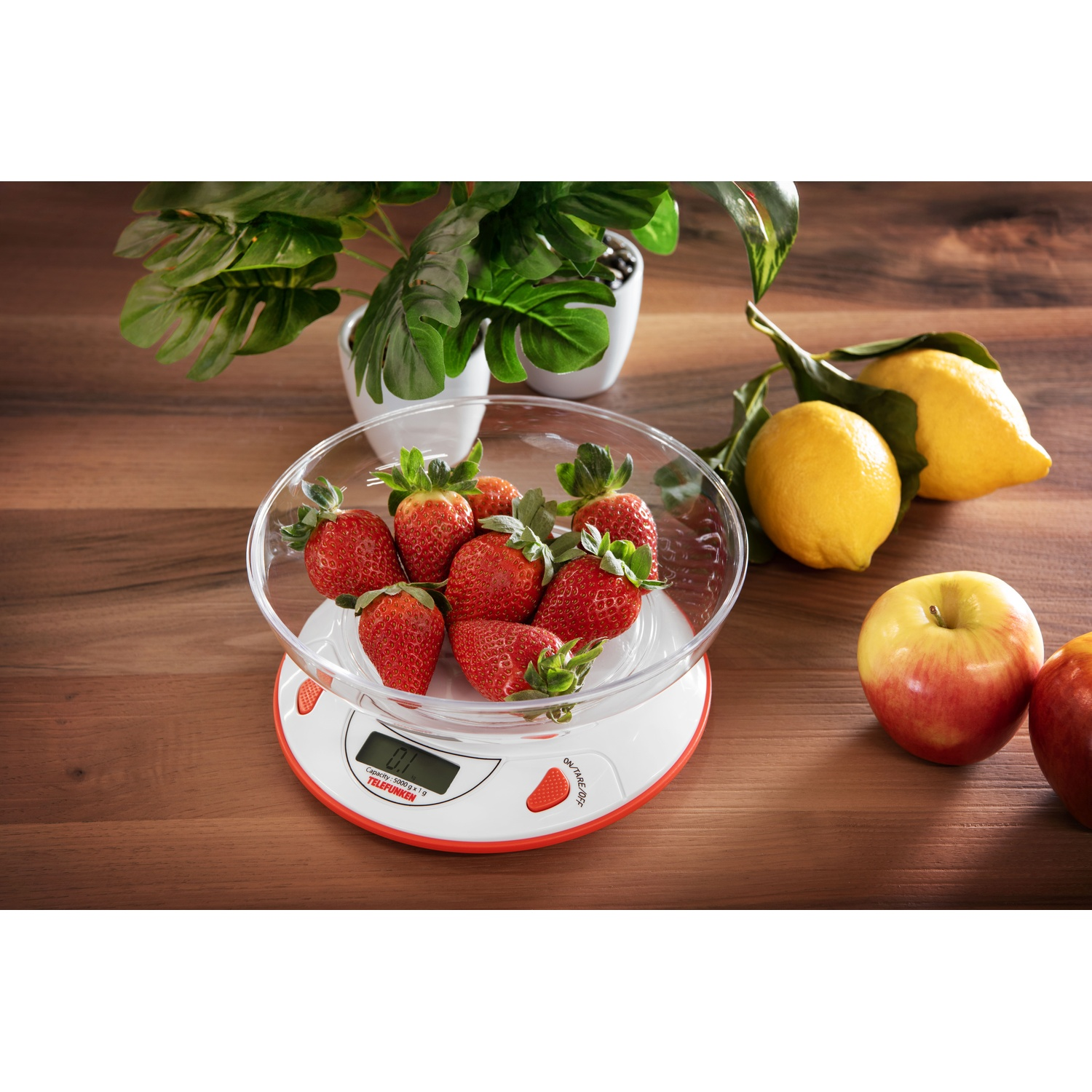Una bilancia con fragole, limoni e una mela su un tavolo di legno con una pianta sullo sfondo.