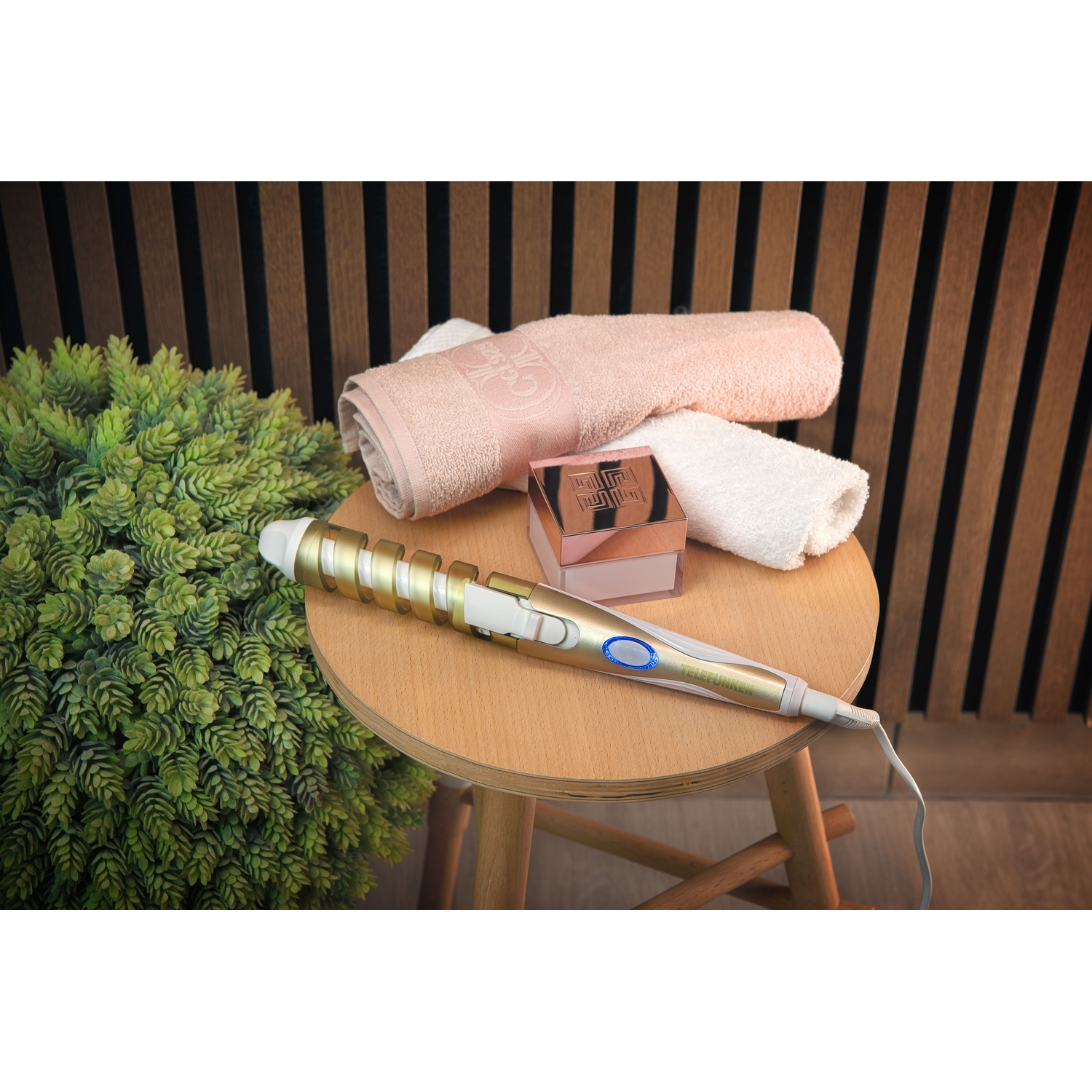 Prodotti e strumenti per lo styling dei capelli su uno sgabello di legno, tra cui un arricciacapelli e un asciugamano rosa.