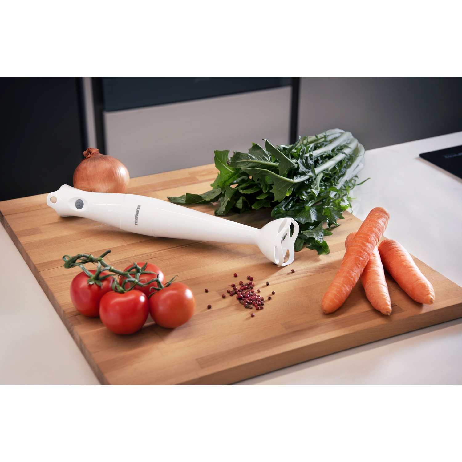Un frullatore a immersione bianco, verdure e spezie su un tagliere di legno.