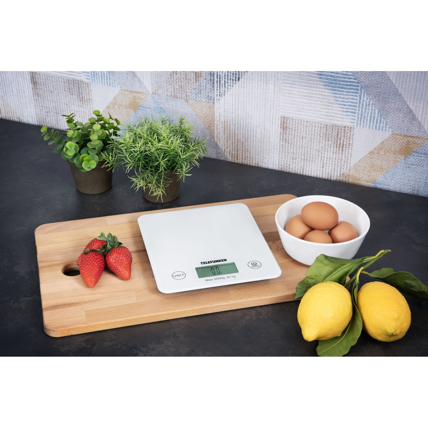 Bilancia da cucina bianca su tagliere. Fragole, uova, limoni e piante presenti.