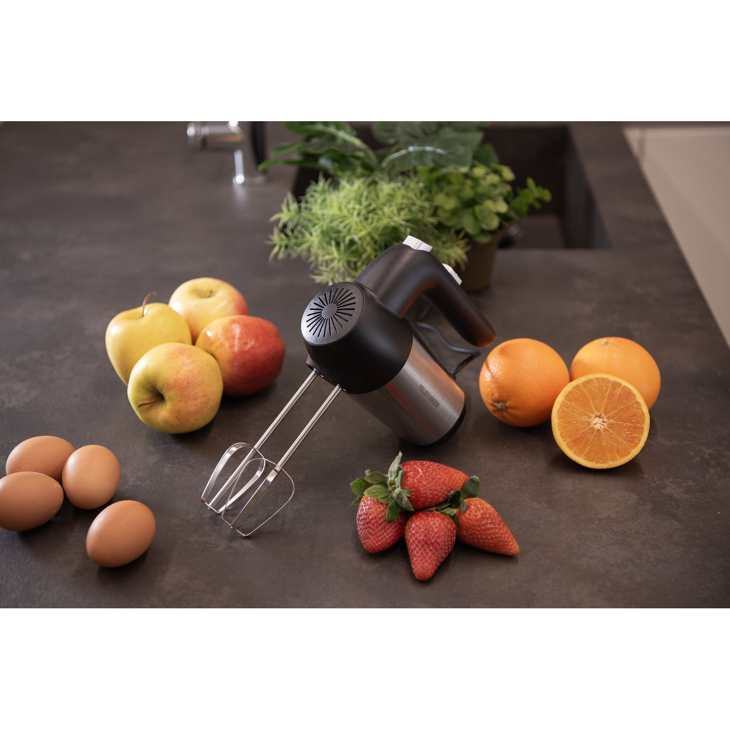 Un frullatore elettrico con frutta e uova su un piano di lavoro. Si vedono mele, arance e fragole.