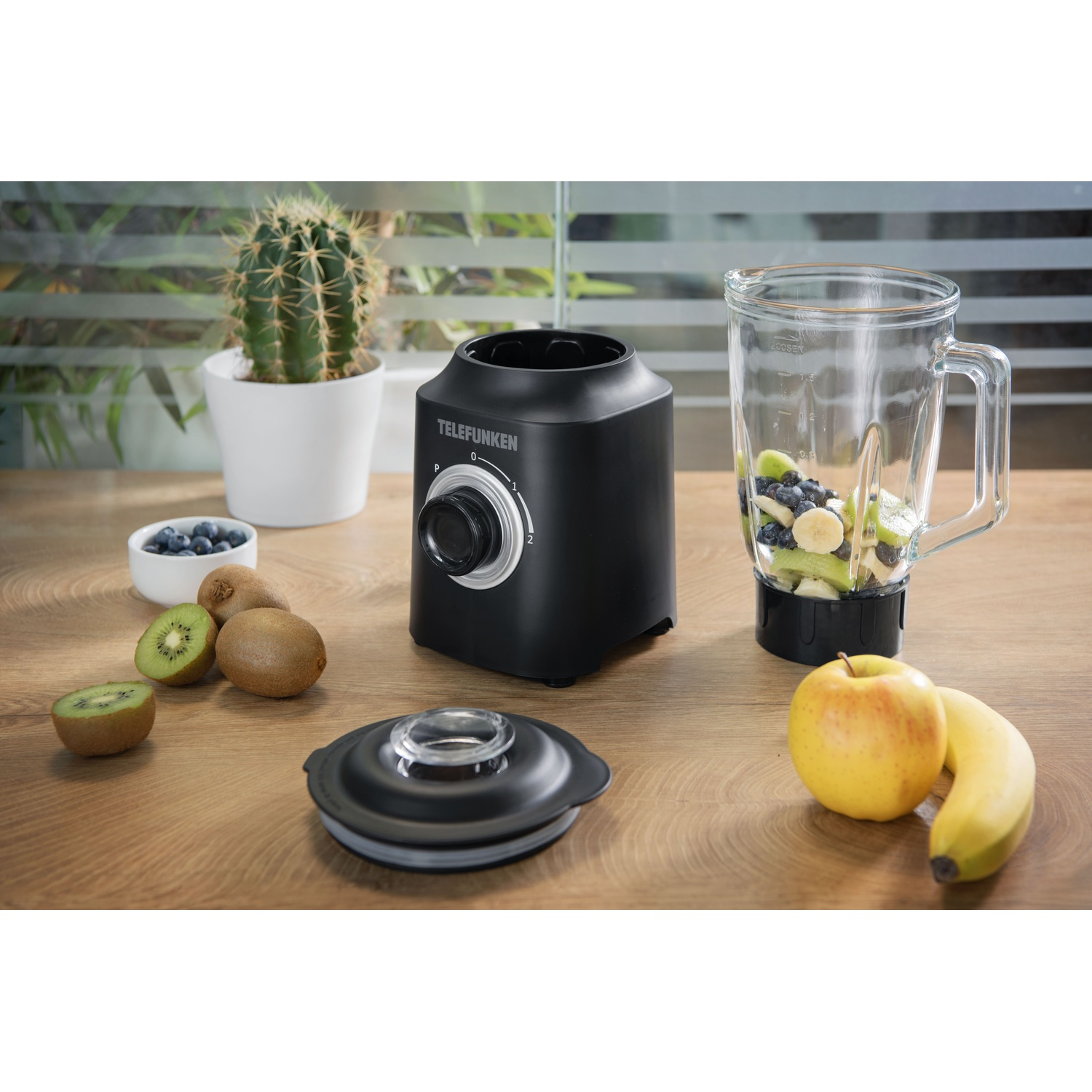 Frullatore con frutta: kiwi, mirtilli, mela e banana, su un tavolo di legno.