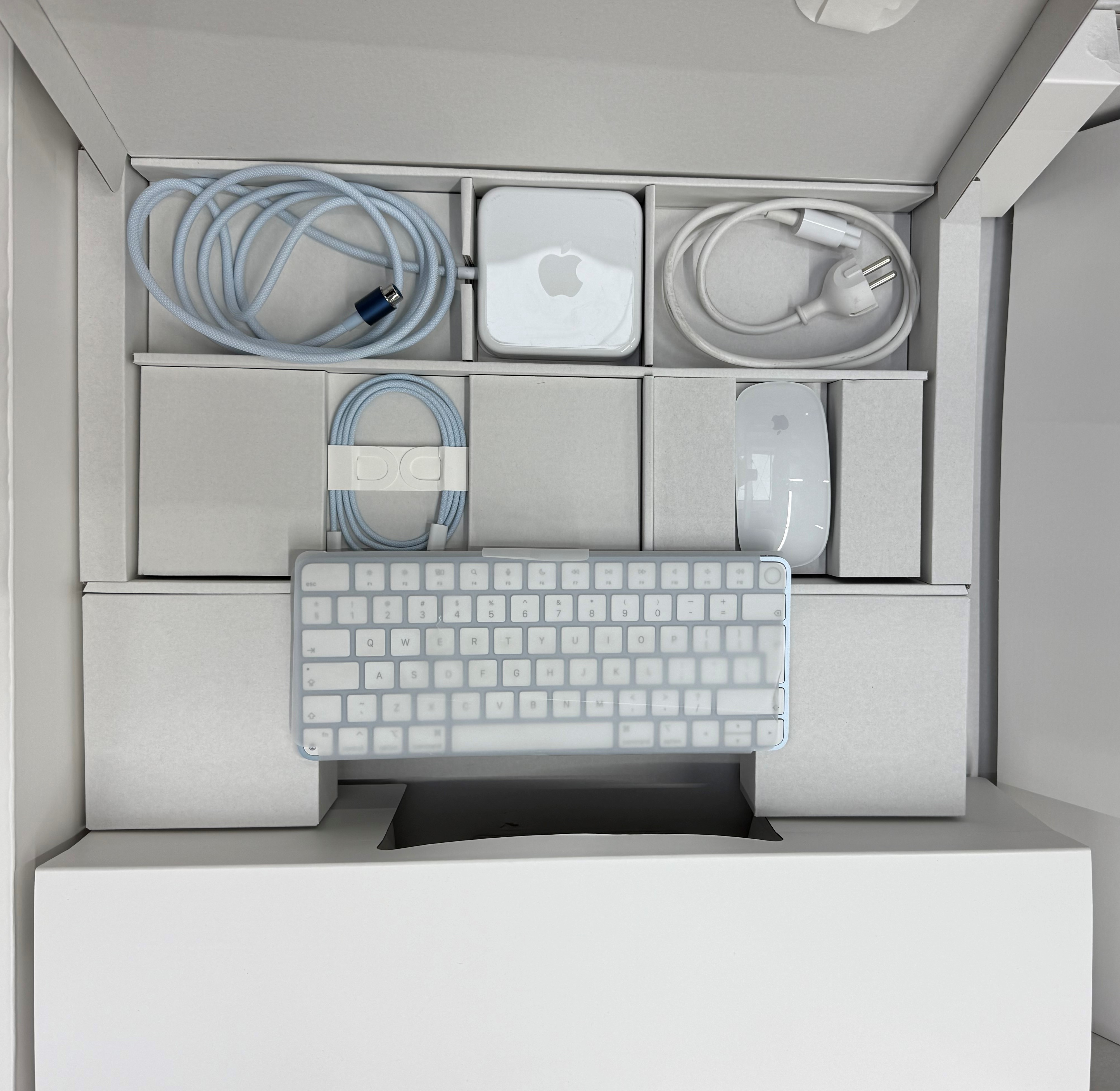 Pudełko wypełnione białą klawiaturą, myszą, kablami i małym komputerem. Wszystkie elementy są białe lub jasnoniebieskie.