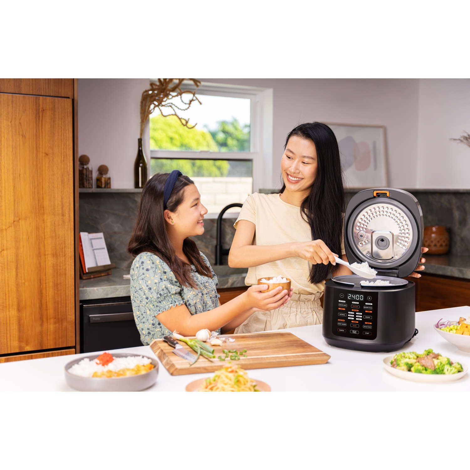 Two women are cooking with a rice cooker. One is serving rice, and the other is holding a bowl.
