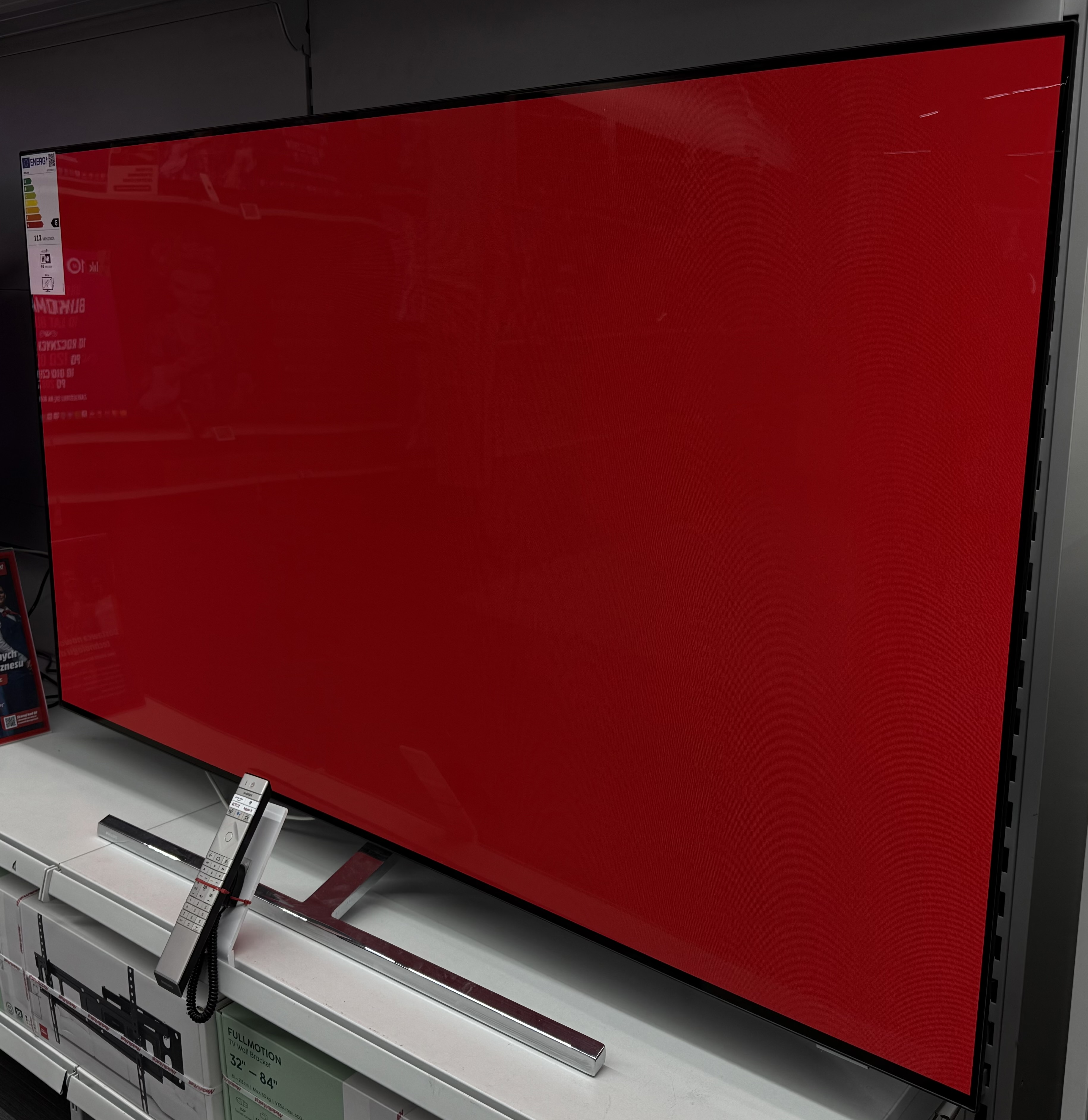 Duży ekran telewizora wyświetlający jednolity czerwony kolor, z pilotem i półką wystawową.