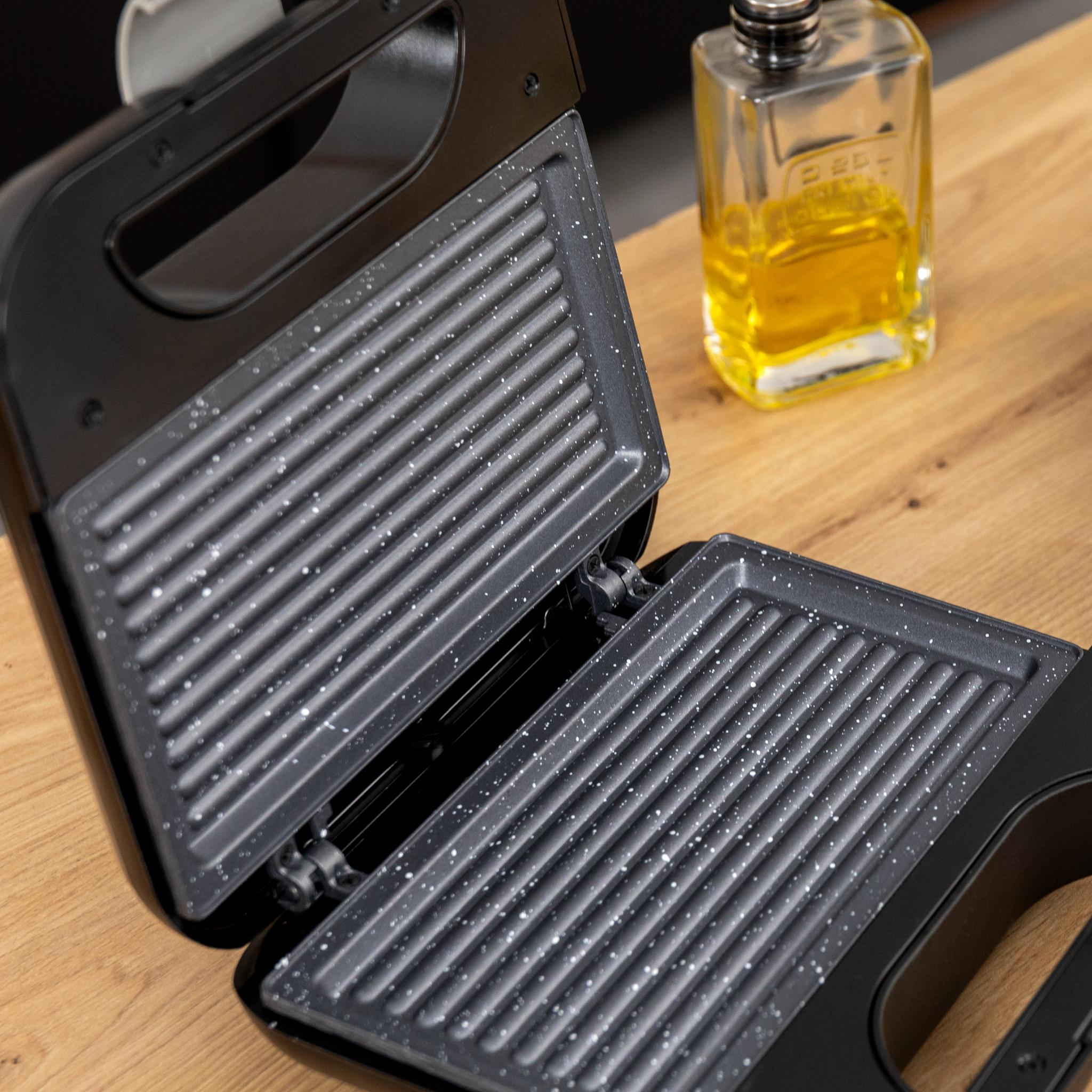 Sandwichera negra abierta con placas texturizadas. Botella de aceite al fondo sobre superficie de madera.