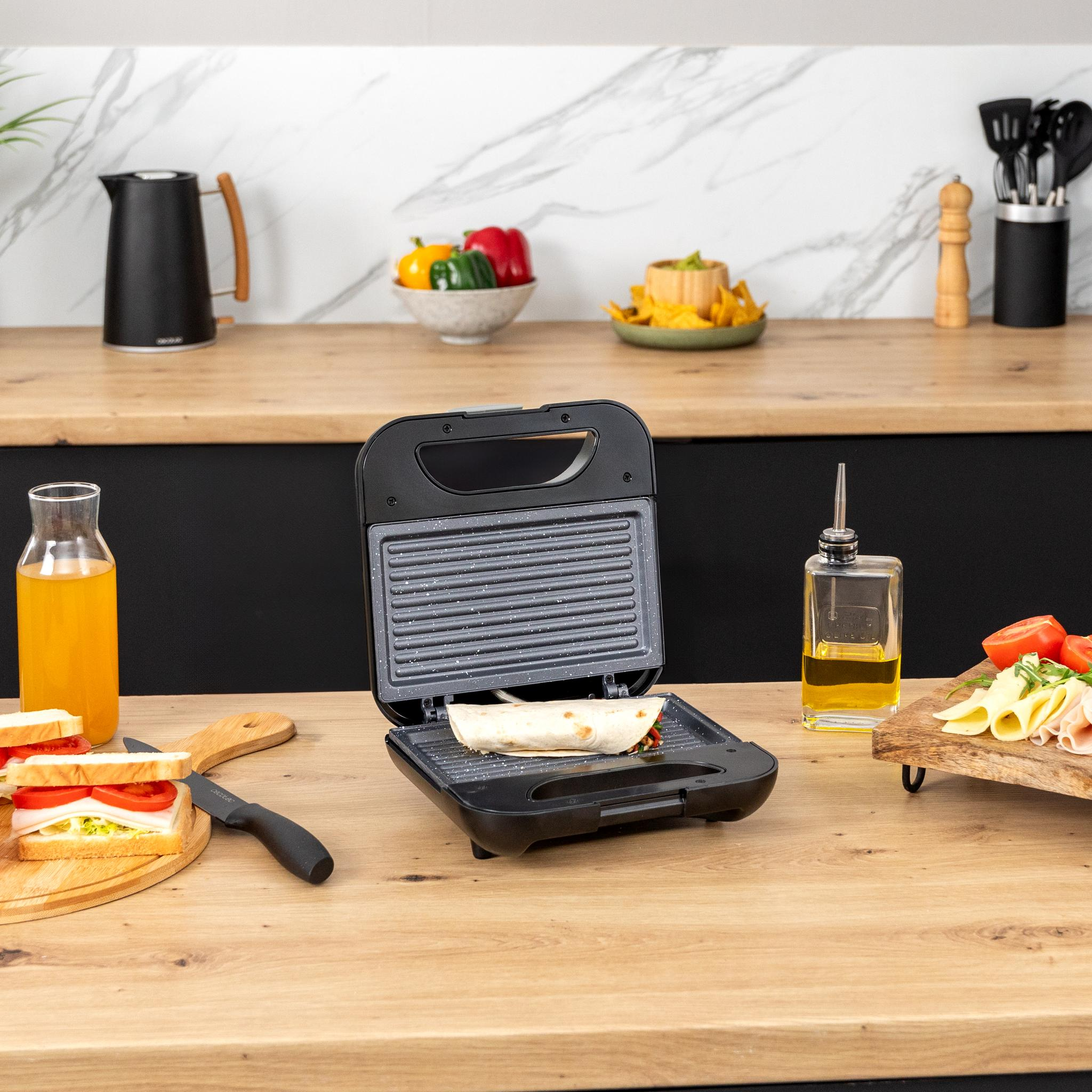 Una sandwichera abierta sobre una superficie de madera. Contiene un sándwich, aceite y varios ingredientes.