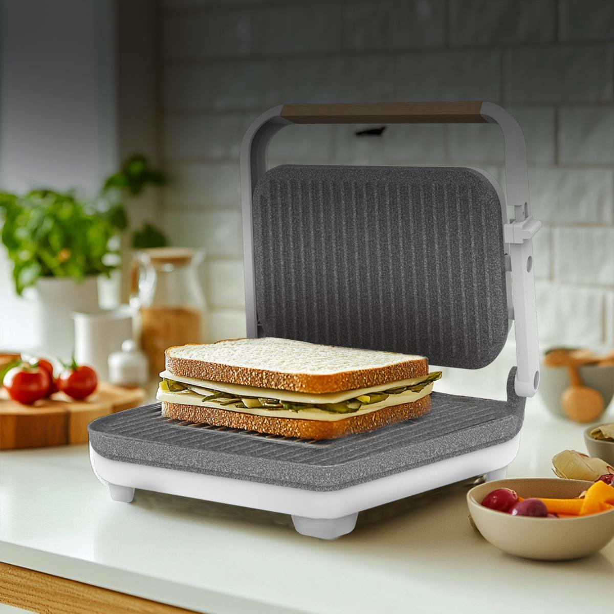 Sándwich en una plancha panini sobre mostrador con tomates y cuencos.