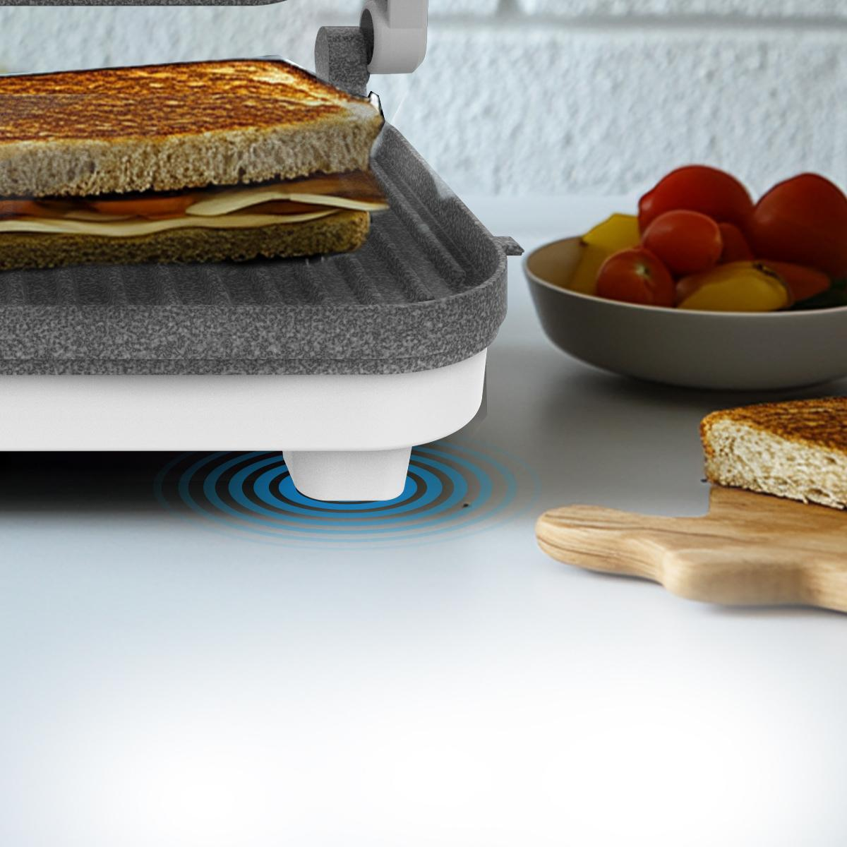Sándwich en una sandwichera. Tomates y una tabla de madera están al lado.