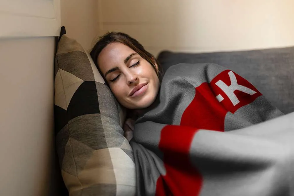 Egy nő alszik a kanapén, betakarózva egy takaróval. Szürke inget visel. Egy párna van mellette.