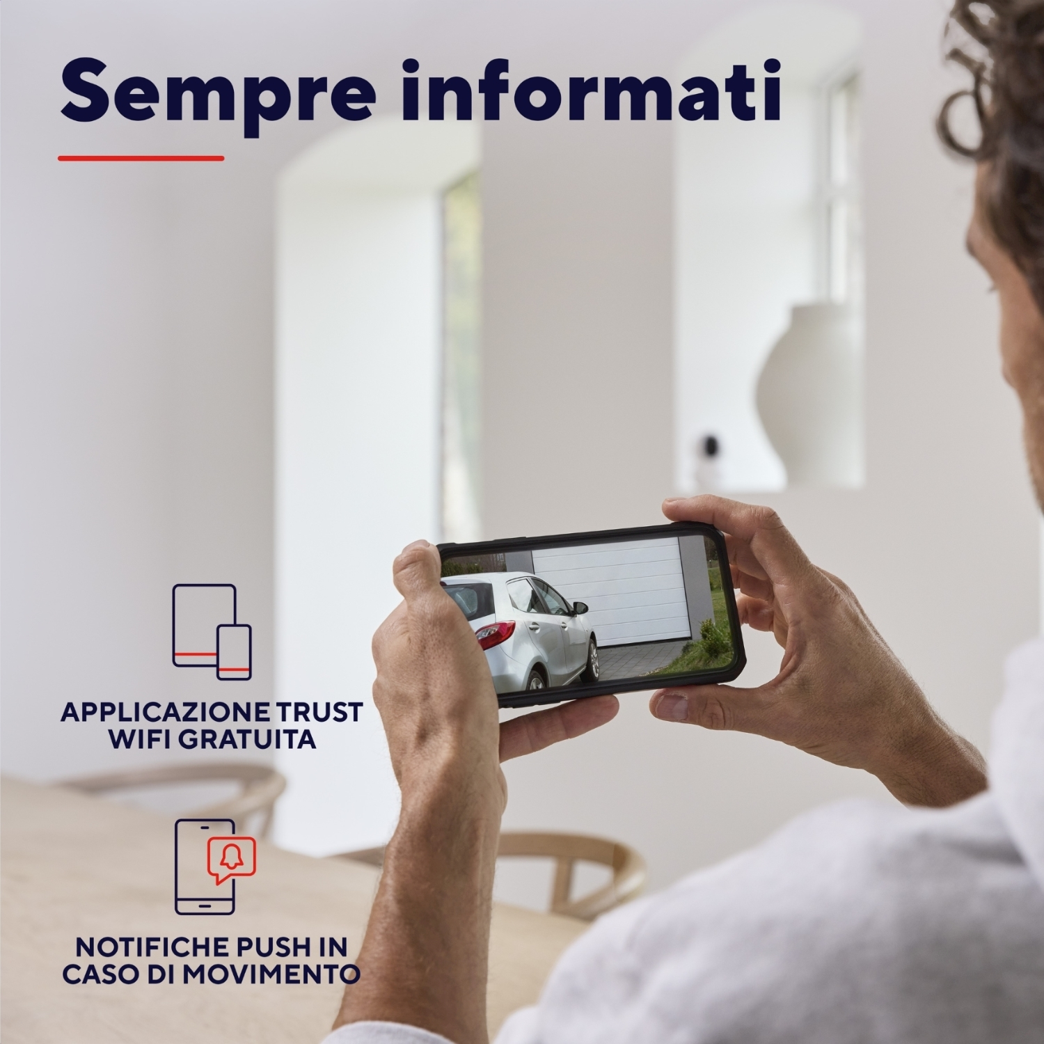Uomo che tiene in mano un telefono che mostra un'auto bianca in un garage. Testo: Sempre informati.