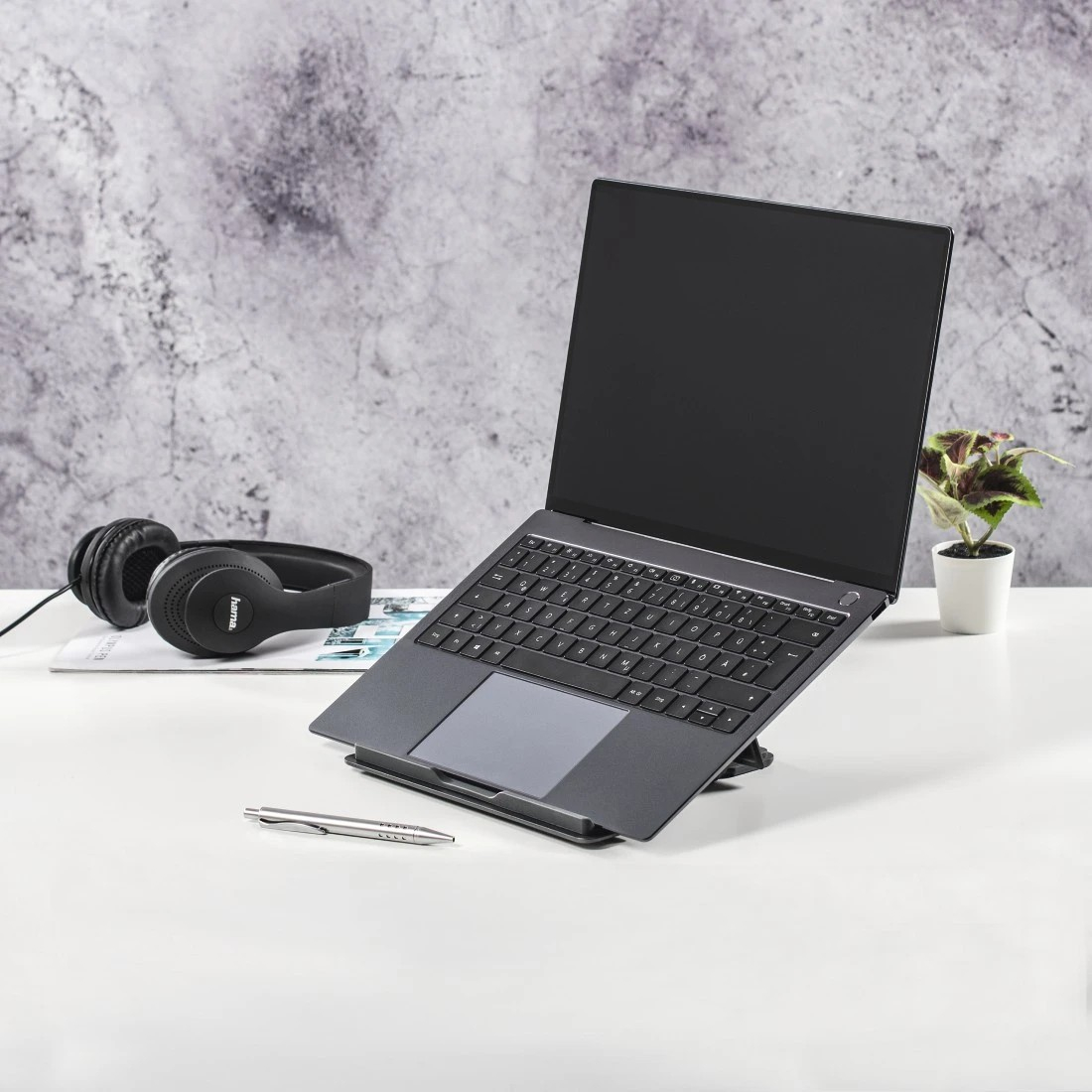 Laptop auf Ständer mit Kopfhörern, Stift und Pflanze auf weißem Schreibtisch. Graue Wand im Hintergrund.