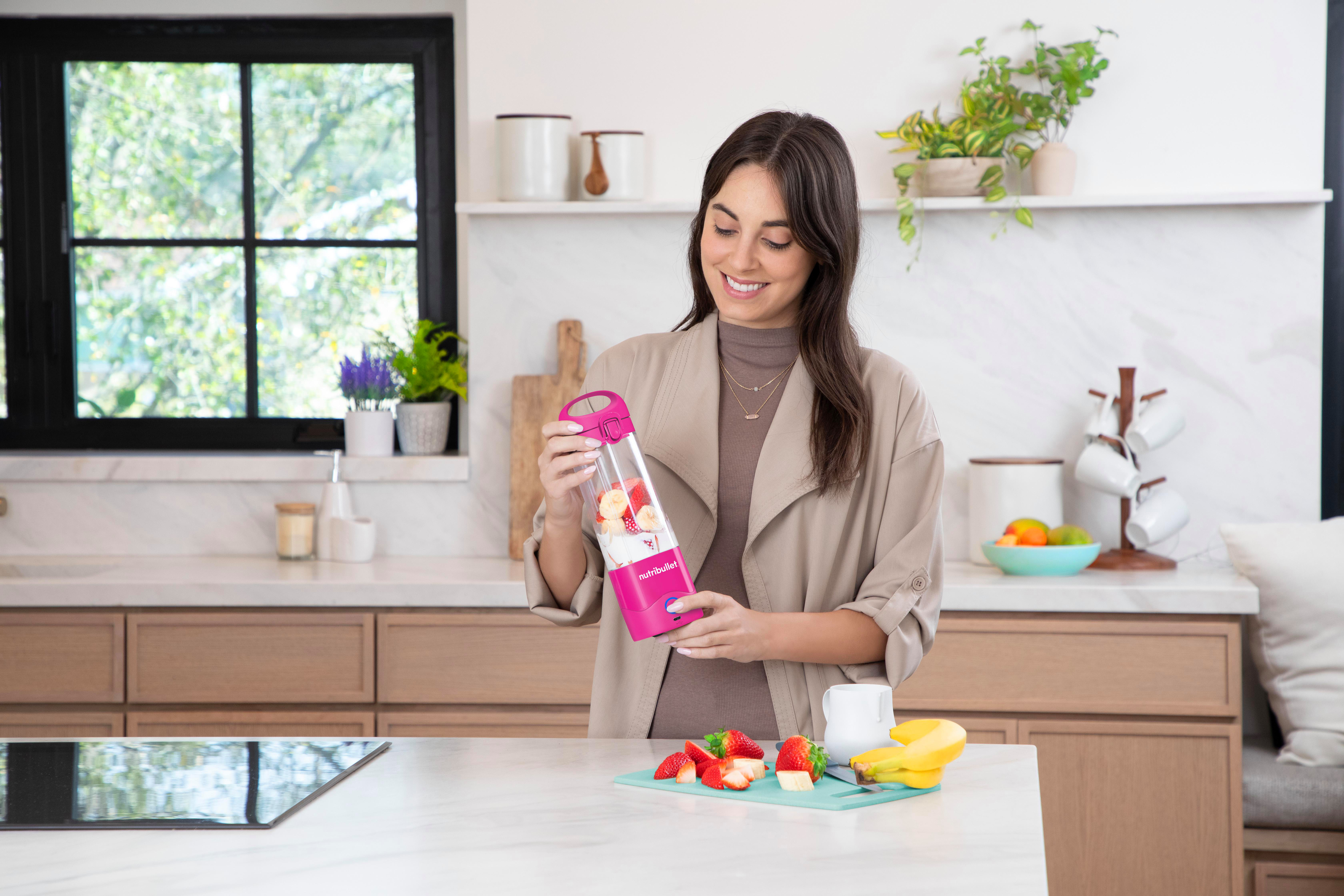 Frau in einer Küche bereitet einen Smoothie zu. Sie hält einen rosa Mixer in der Nähe von Früchten.