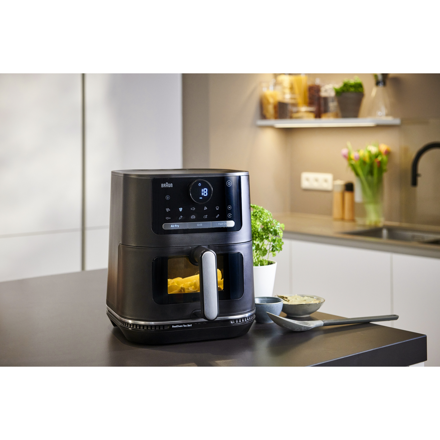 Une friteuse à air sur un comptoir avec de la nourriture à l'intérieur, près d'une plante et d'articles de cuisine.
