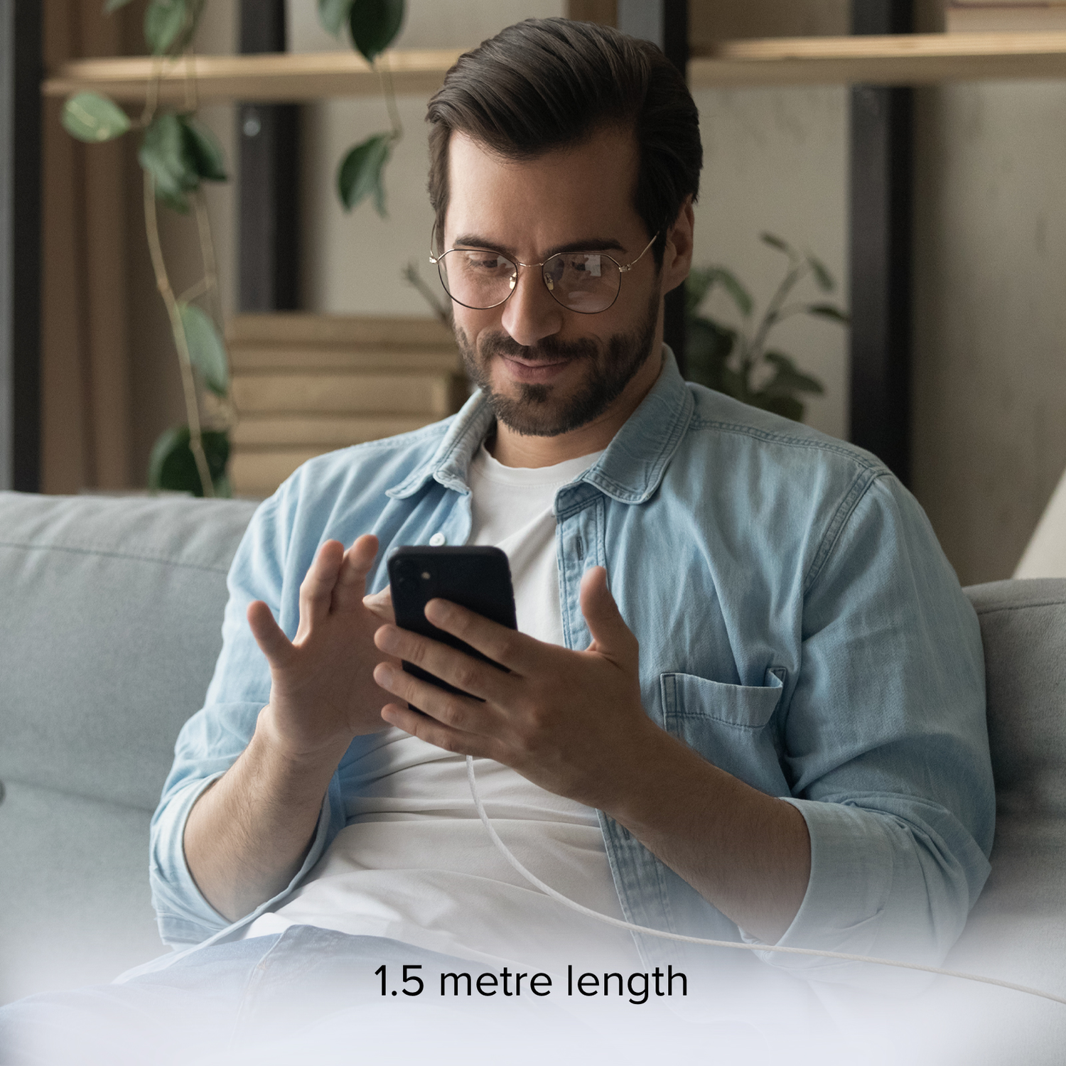 A man with glasses sits on a sofa looking at a phone, 1.5-meter white charging cable.