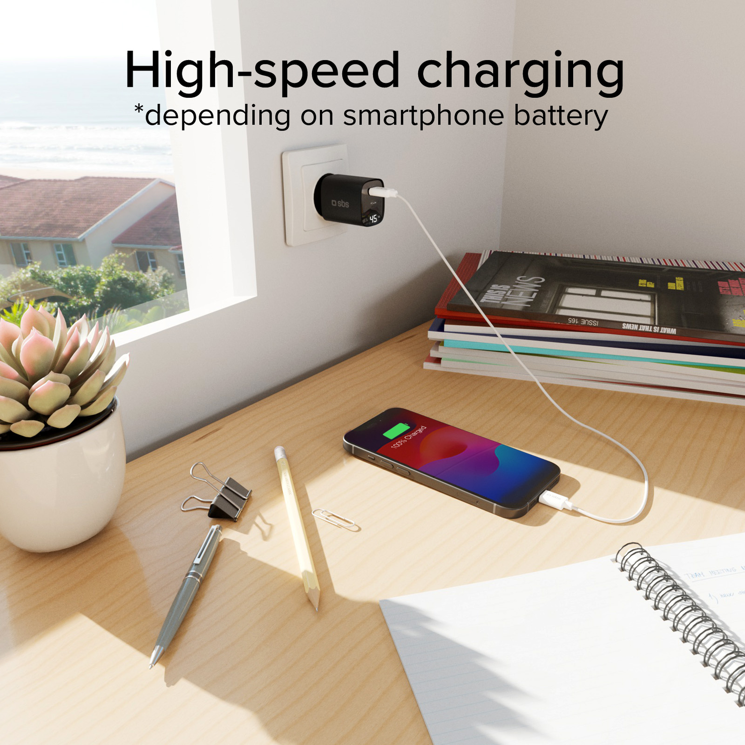 Smartphone en charge avec un câble blanc, branché à une prise murale. Livres et stylo.