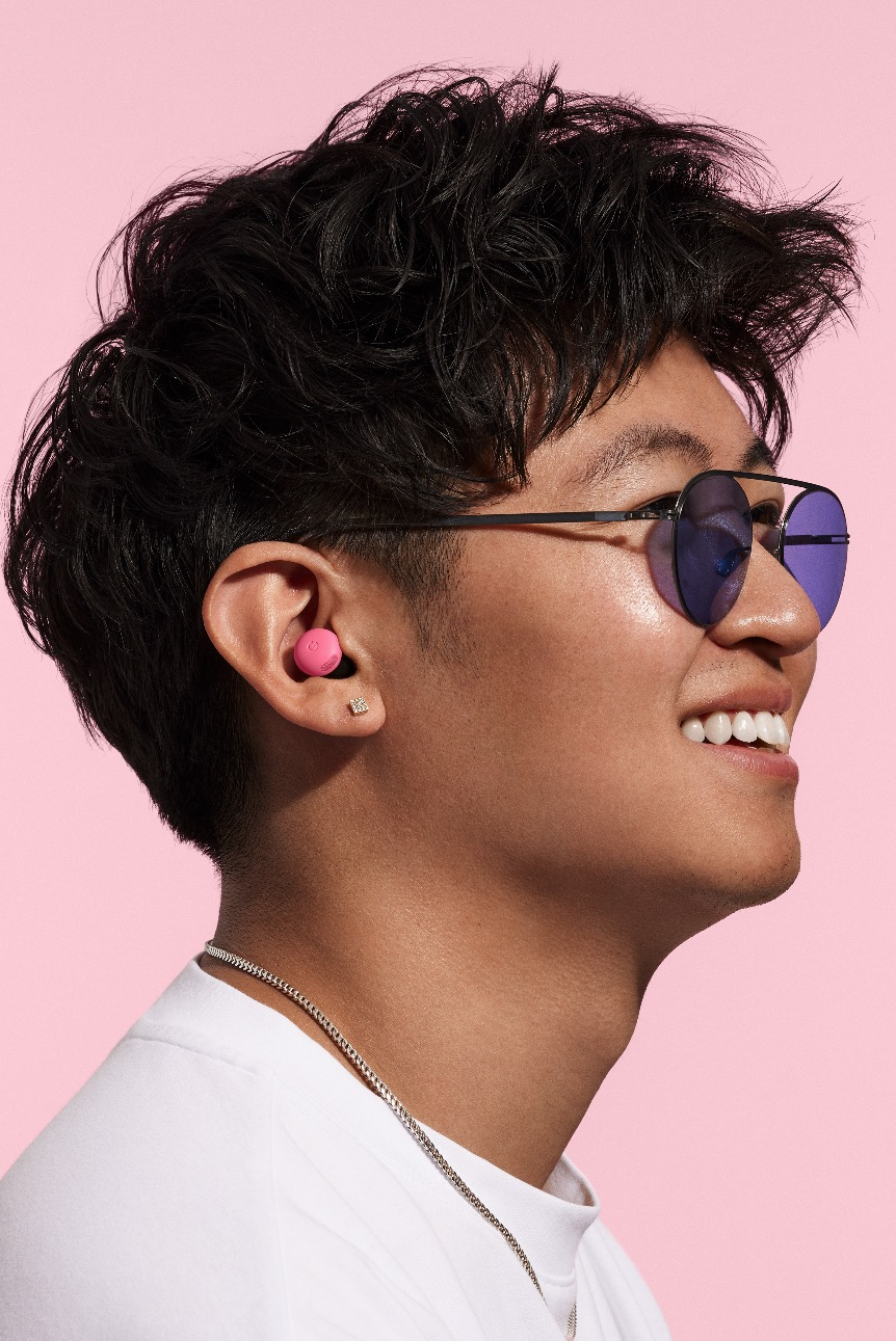 Ein junger Mann mit rosa Ohrhörern, Sonnenbrille. Lächelt vor rosa Hintergrund.