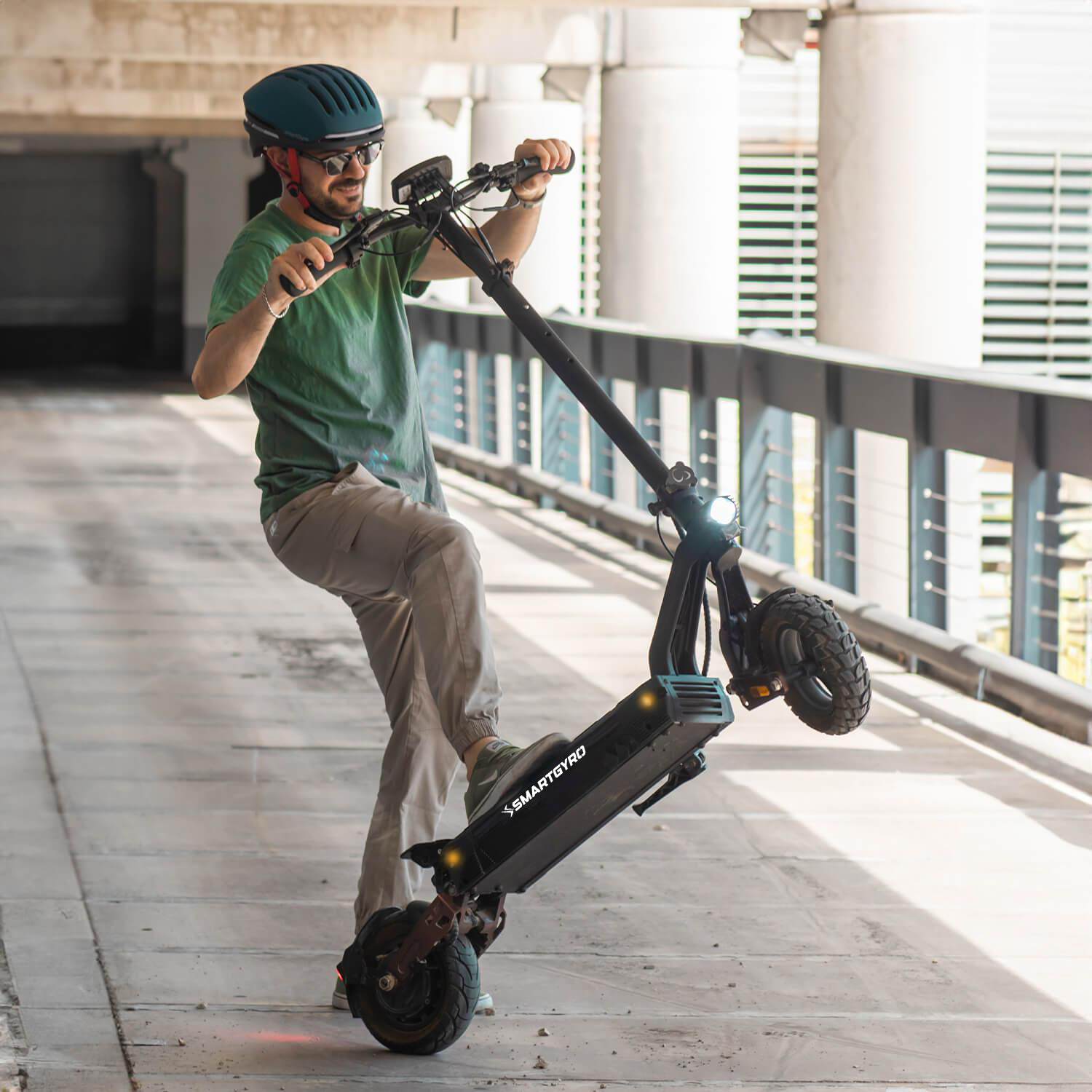 Un hombre se equilibra en un patinete eléctrico. Lleva una camisa verde y está en un entorno urbano. El patinete es negro y gris.
