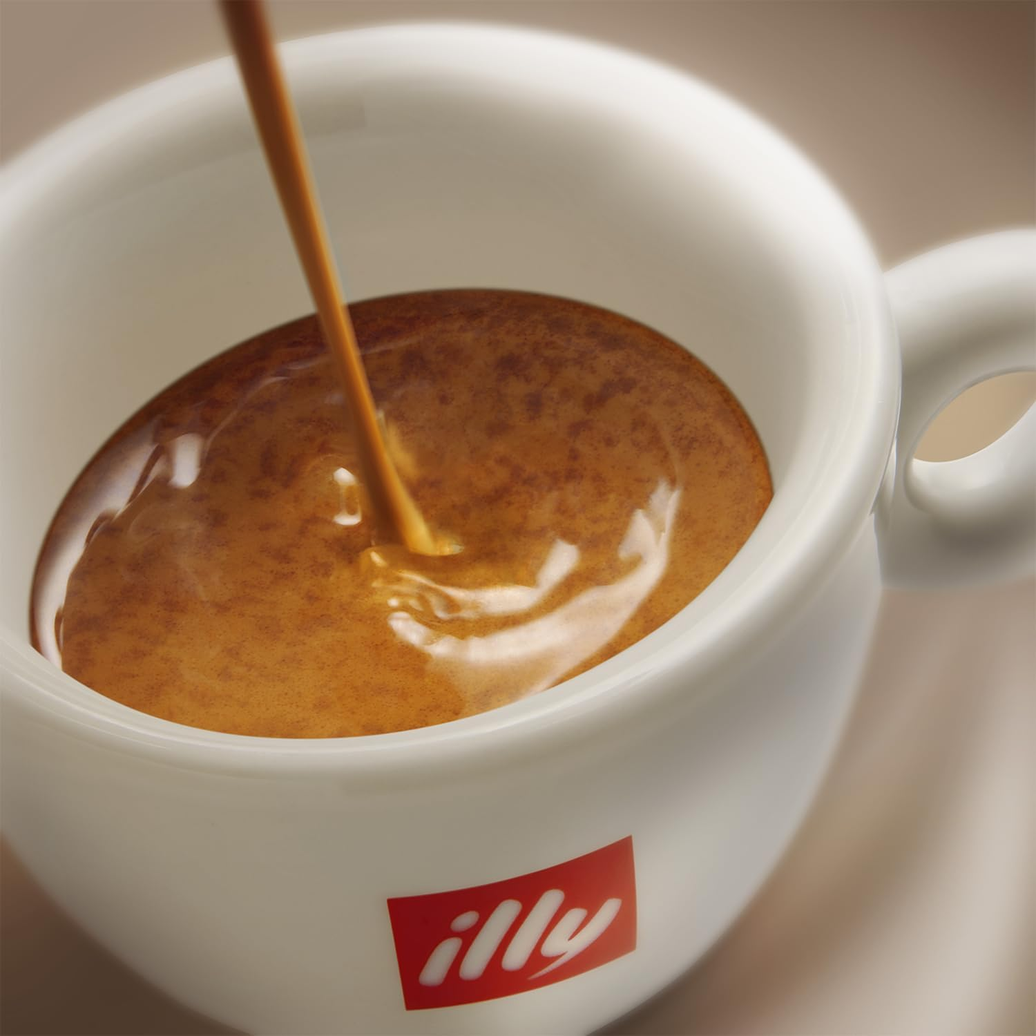 Café vertiéndose en una taza blanca con el logo Illy. El café es marrón y la taza es blanca.