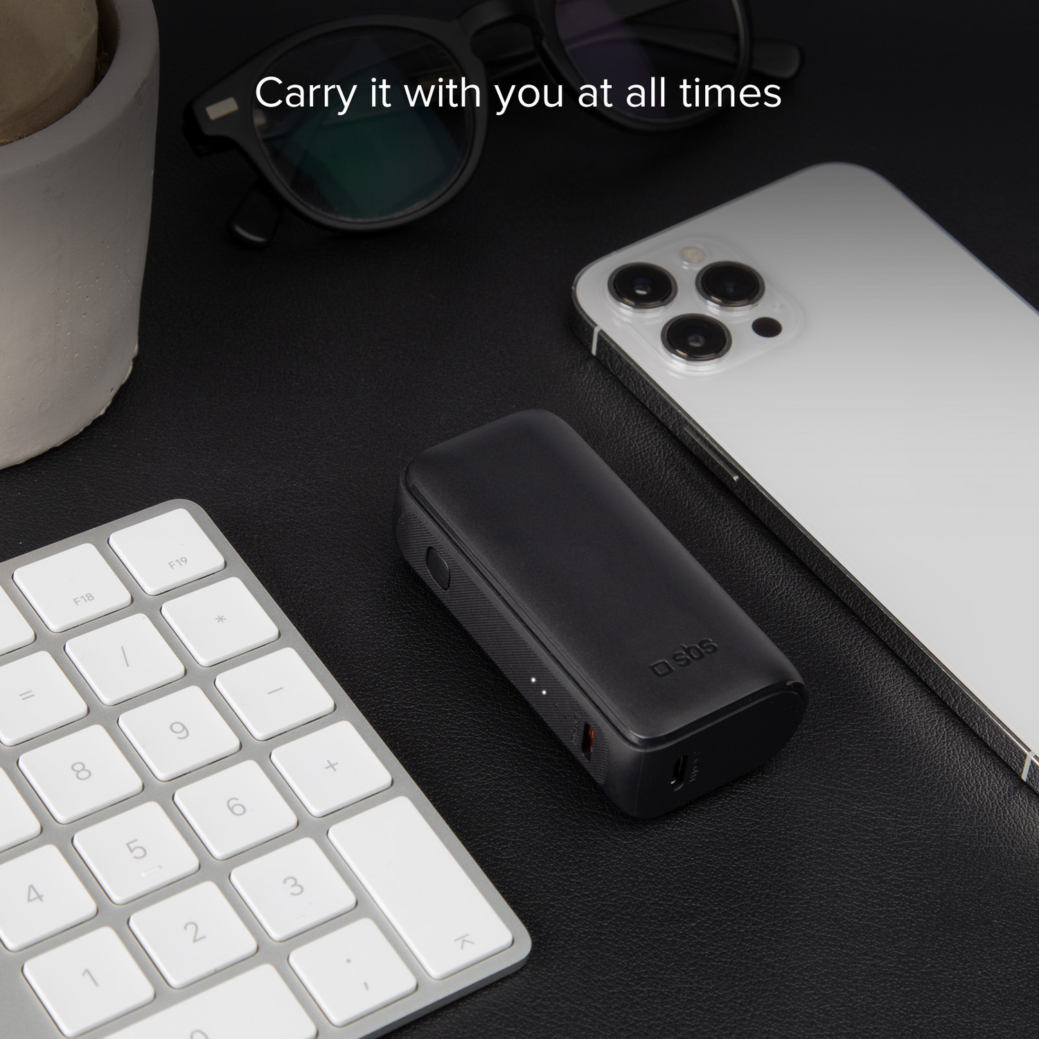 Black portable charger and white smartphone on a table with a keyboard and glasses.