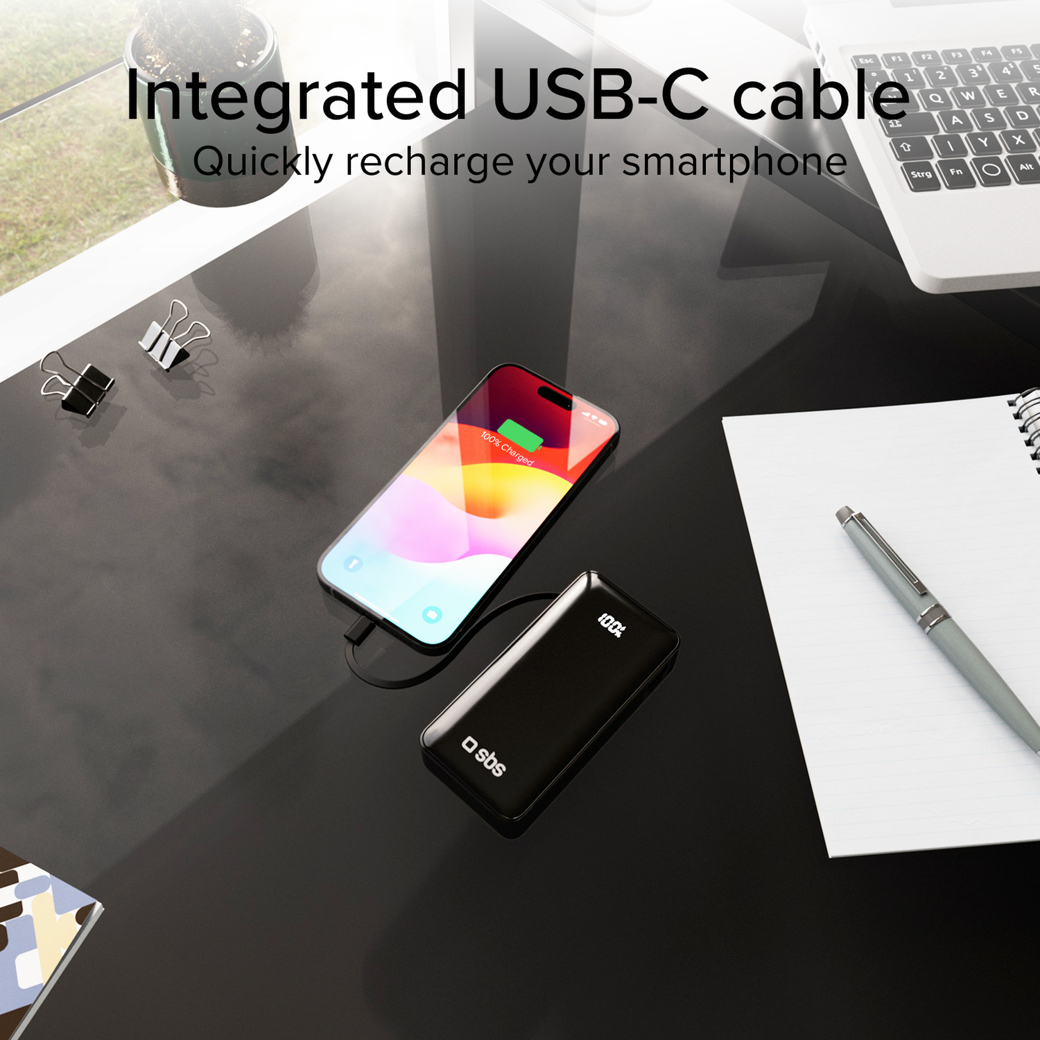 Black power bank charging a phone with 100% charged on a desk with a laptop and pen.