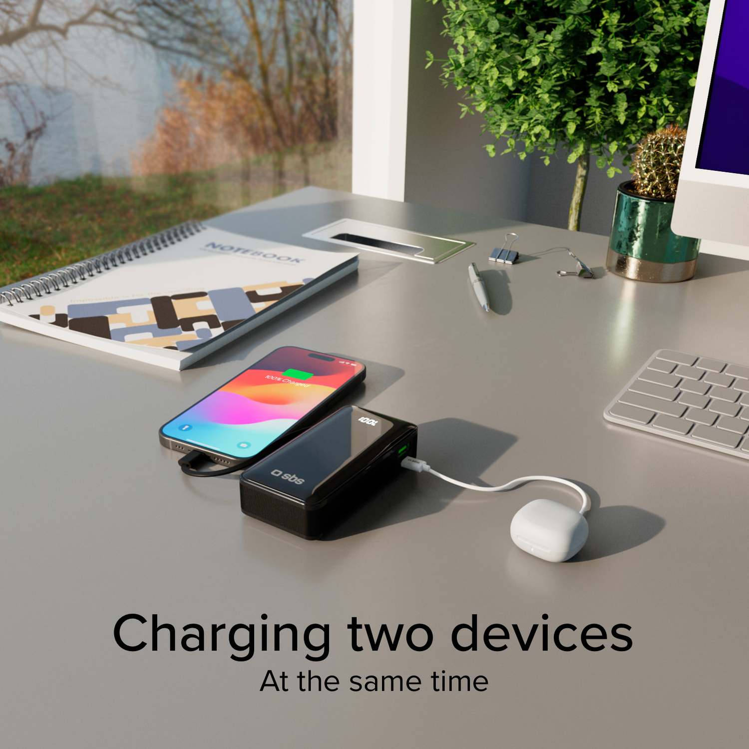 Two devices charging: phone and power bank on a desk with a notebook and a small plant.