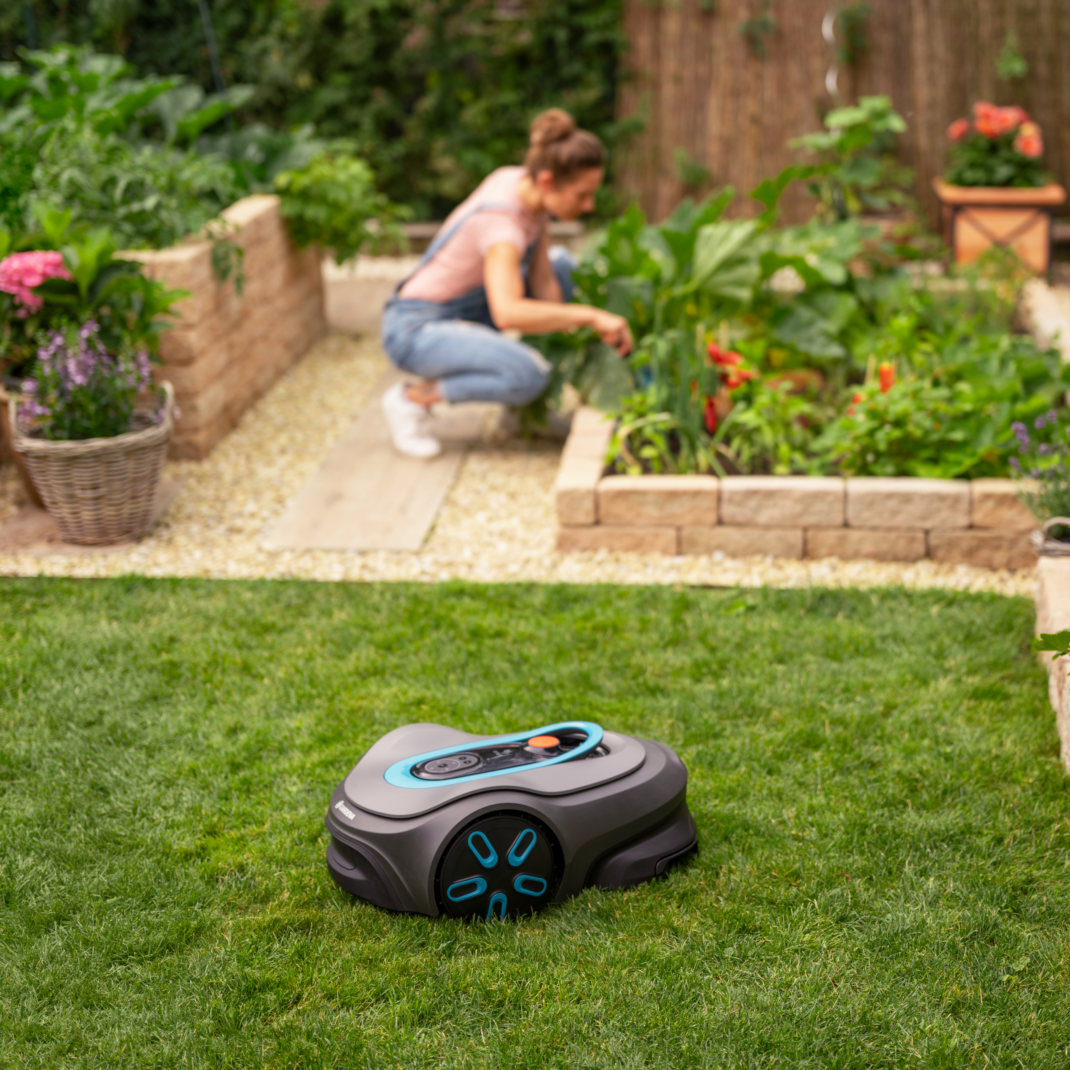 Mähroboter auf grünem Gras mit einer Frau, die im Hintergrund einen Garten pflegt.