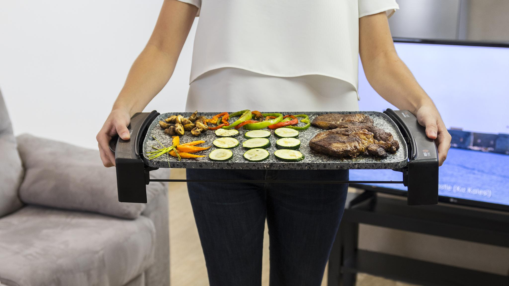 Persona sosteniendo parrilla con verduras y carne cocinadas. Fondo: salón con TV.